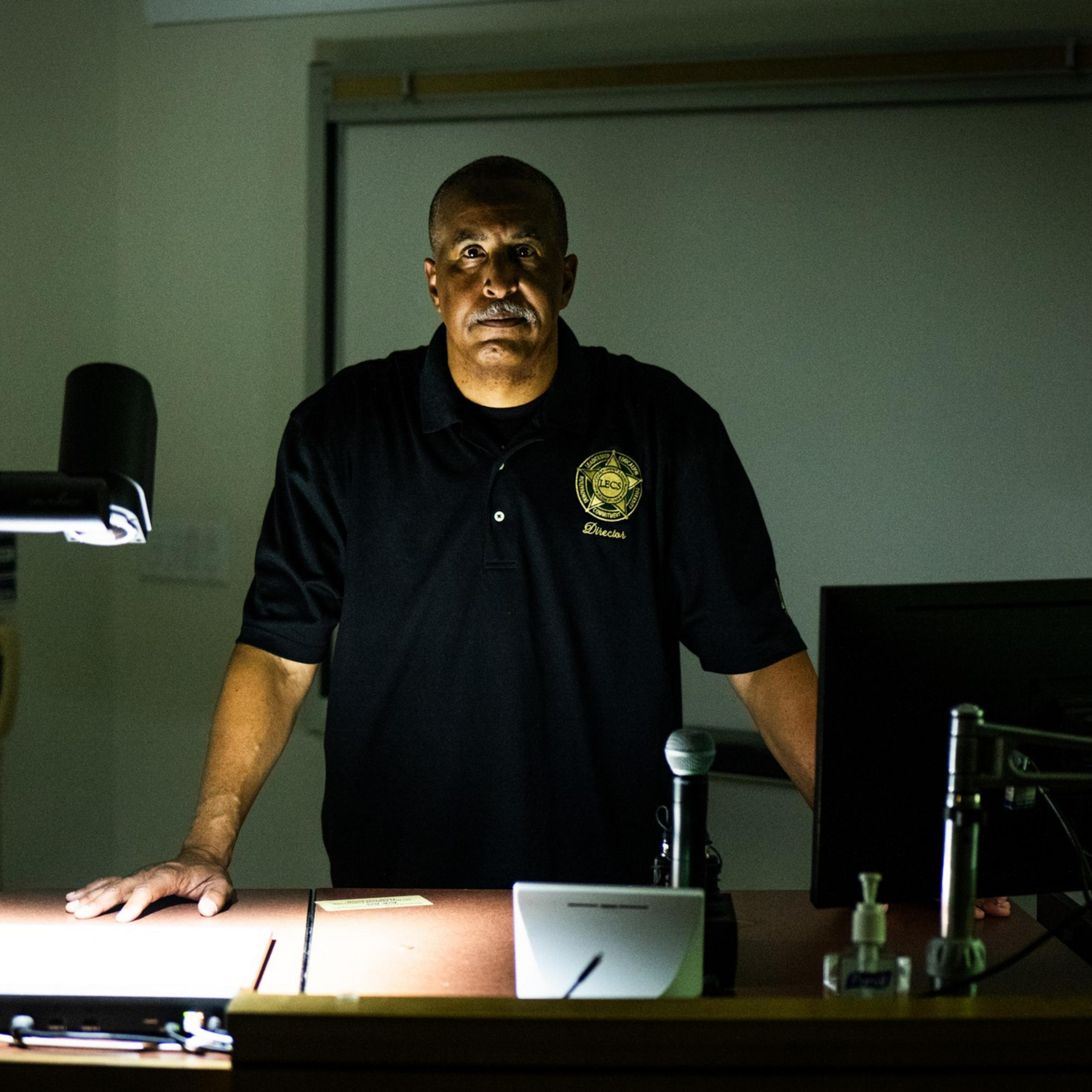 A man in a dark polo shirt stands at a lectern with a computer and a document camera, lit dramatically from above, against a dimly lit classroom backdrop.