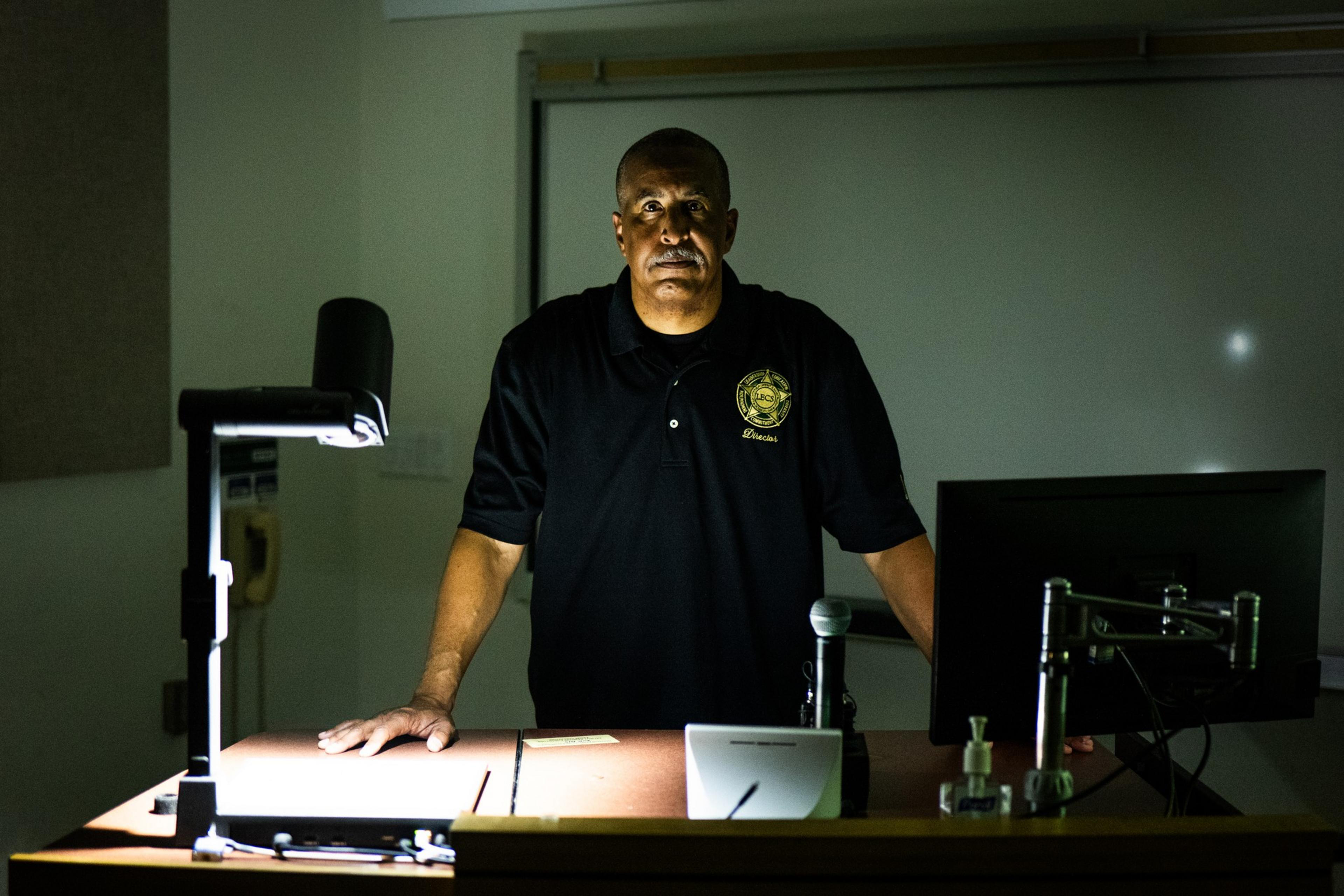 A man in a dark polo shirt stands at a lectern with a computer and a document camera, lit dramatically from above, against a dimly lit classroom backdrop.