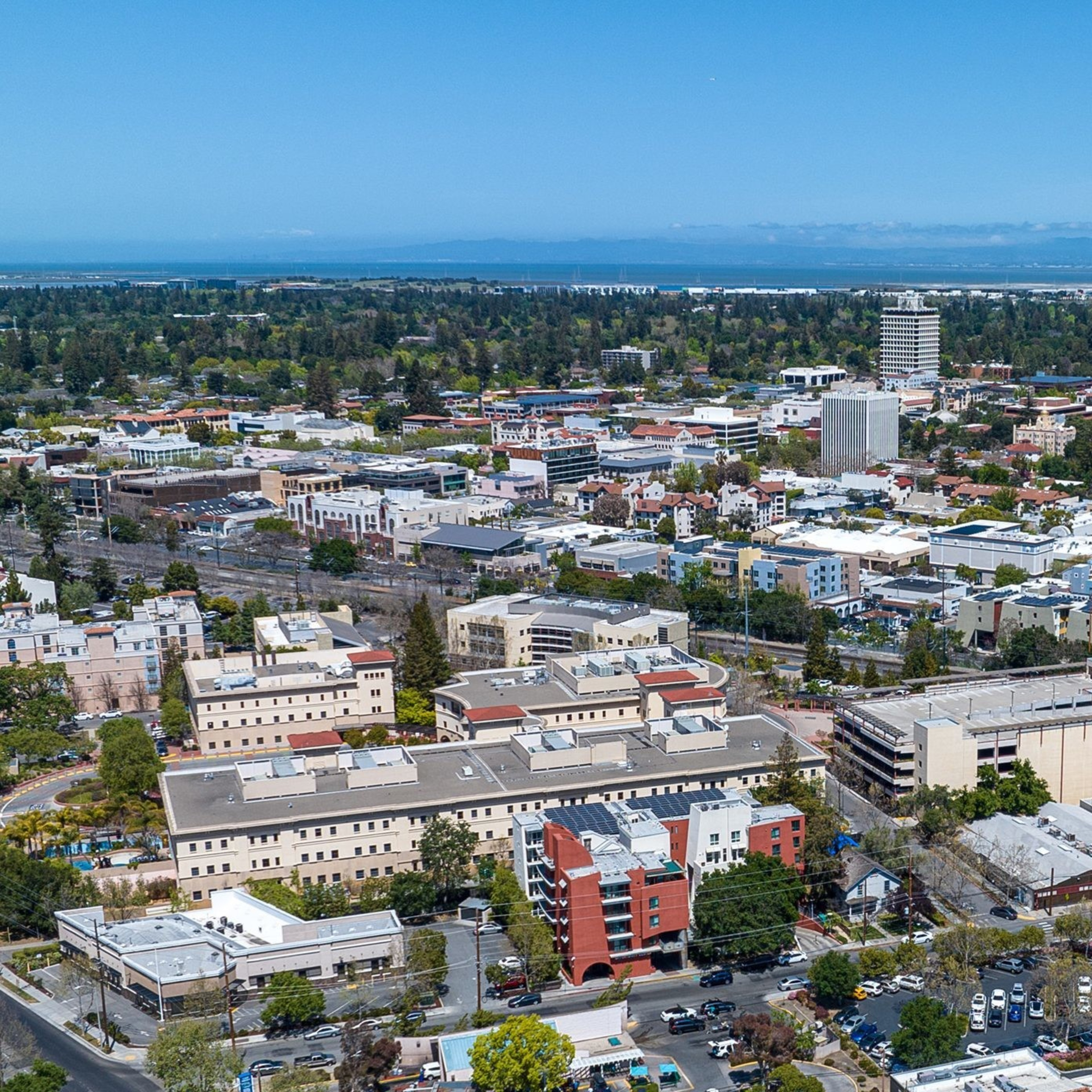 The image shows a cityscape with a mix of low-rise and mid-rise buildings surrounded by trees. In the background, there are distant hills under a clear blue sky.