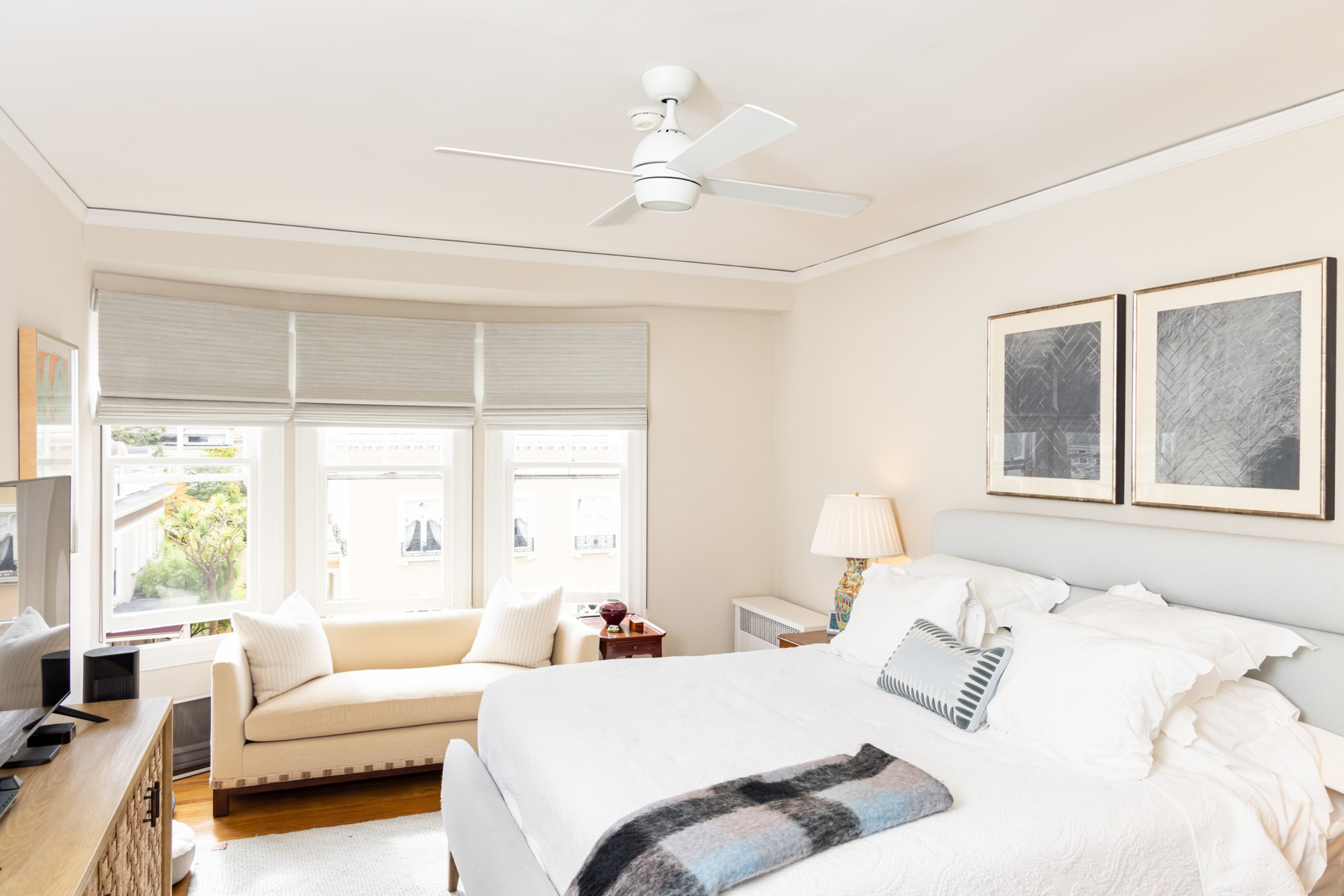 A bright bedroom features a large bed with white bedding, two framed pictures above the headboard, a ceiling fan, a cream loveseat by windows, and a wooden dresser with a TV.