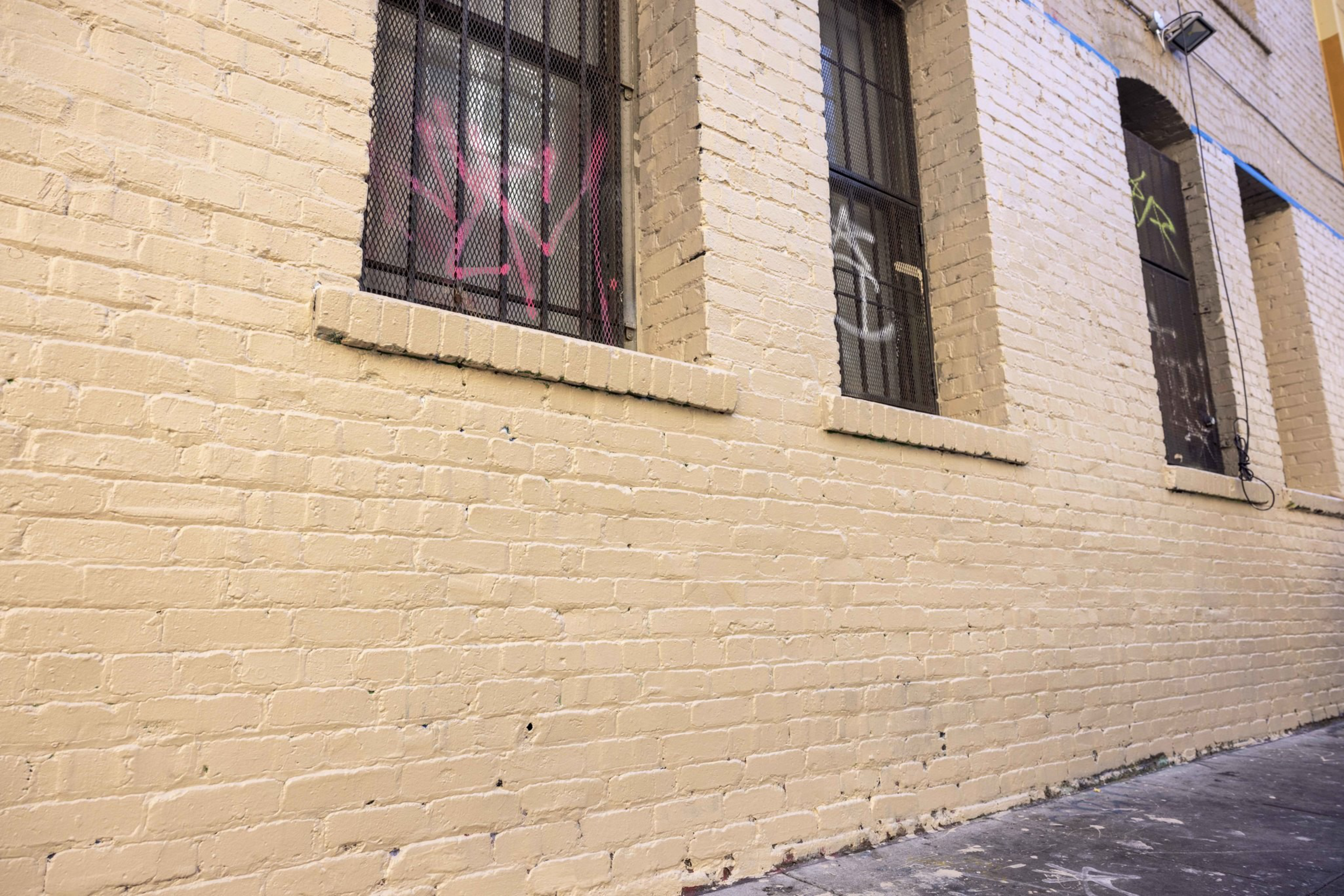 A beige brick wall with barred windows featuring pink, white, and green graffiti. The ground is gray, and there's noticeable wear on the surface.