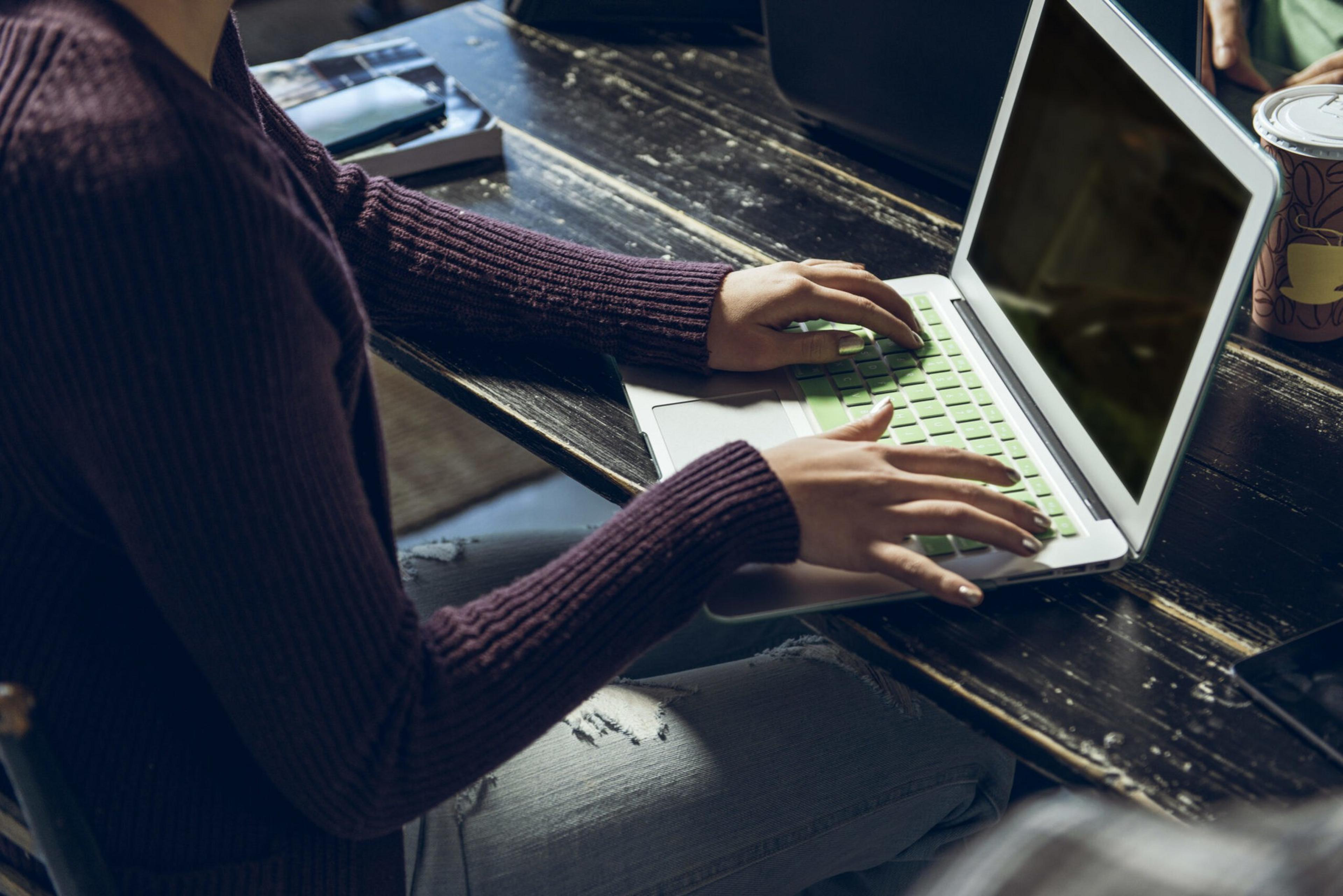 A person wearing a dark purple sweater types on a laptop with a green keyboard cover, sitting at a worn wooden table with a coffee cup nearby.