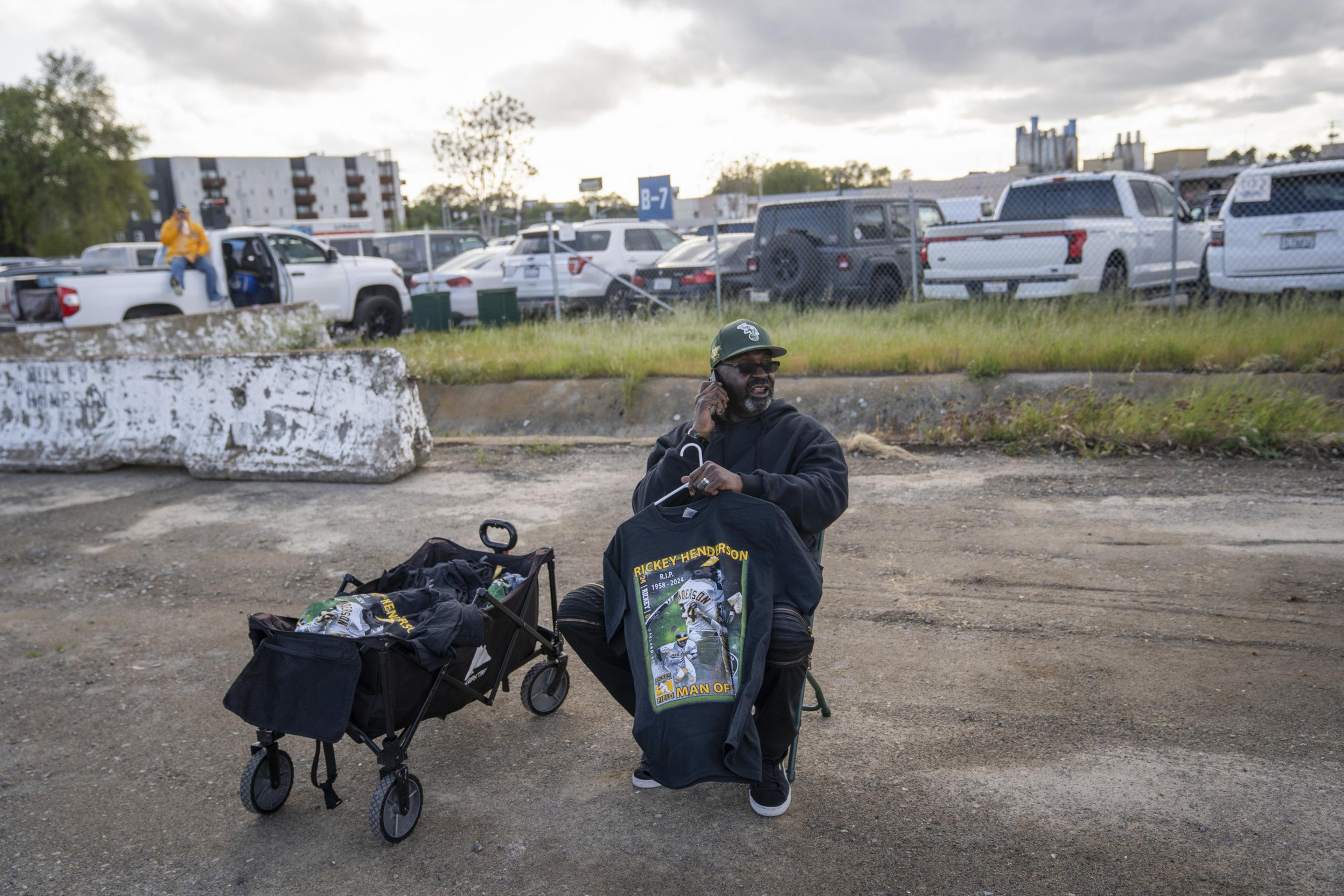 A man sits on a chair in a parking lot, holding up a black T-shirt with a design. A cart beside him holds more shirts, and there are cars parked in the background.