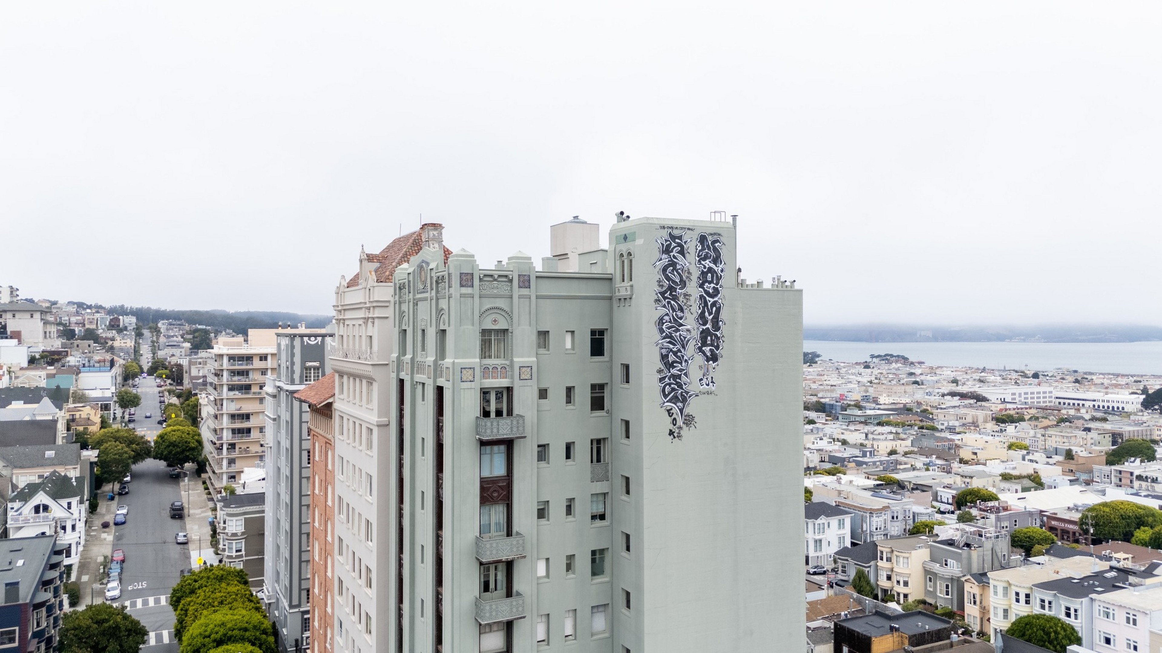 A tall, pale green building with decorative architecture features a large vertical black and white mural on its side, overlooking a cityscape and waterfront under an overcast sky.