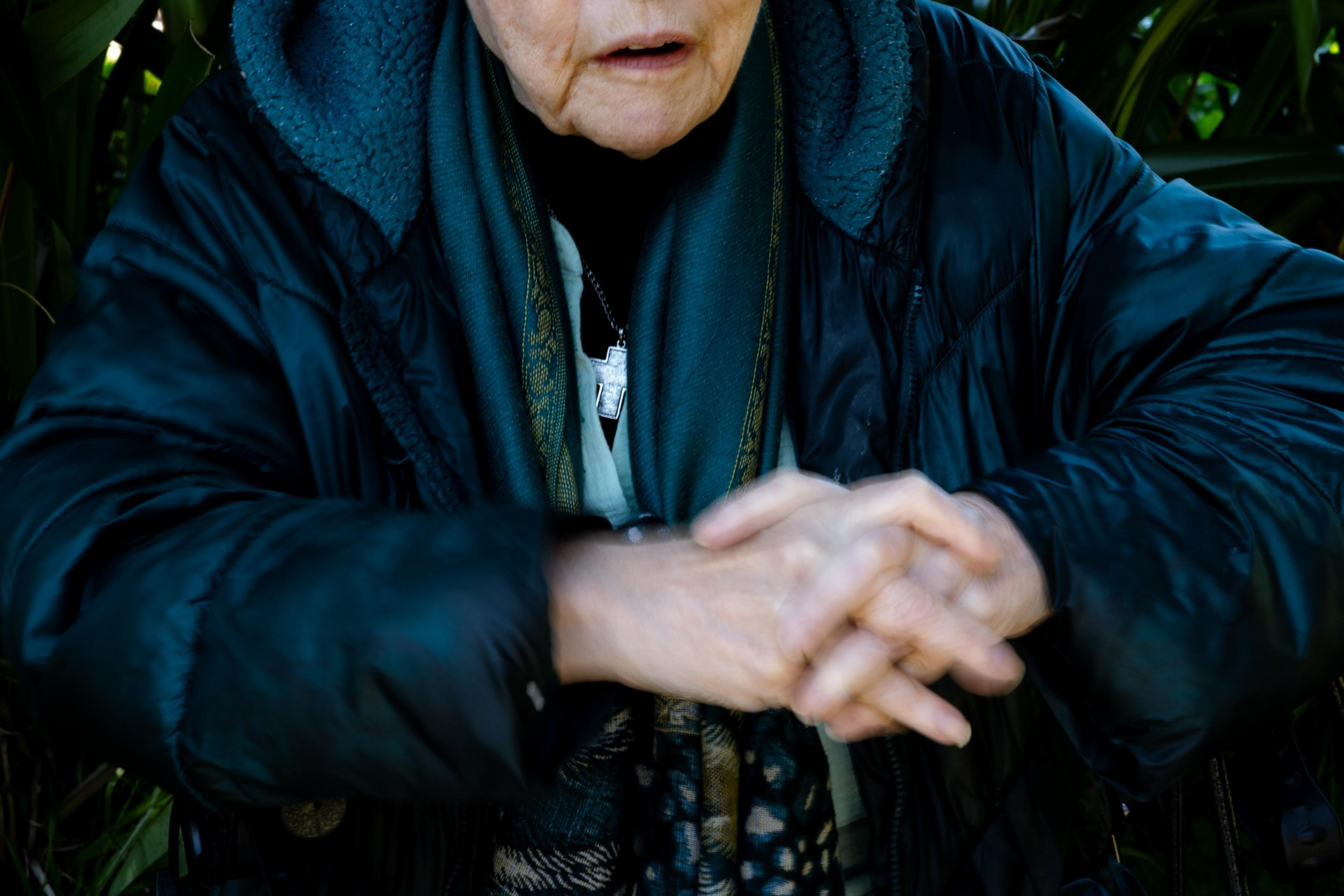 An older individual in a heavy black coat and teal scarf clasps hands together. A silver cross necklace is visible against the dark background of the jacket.