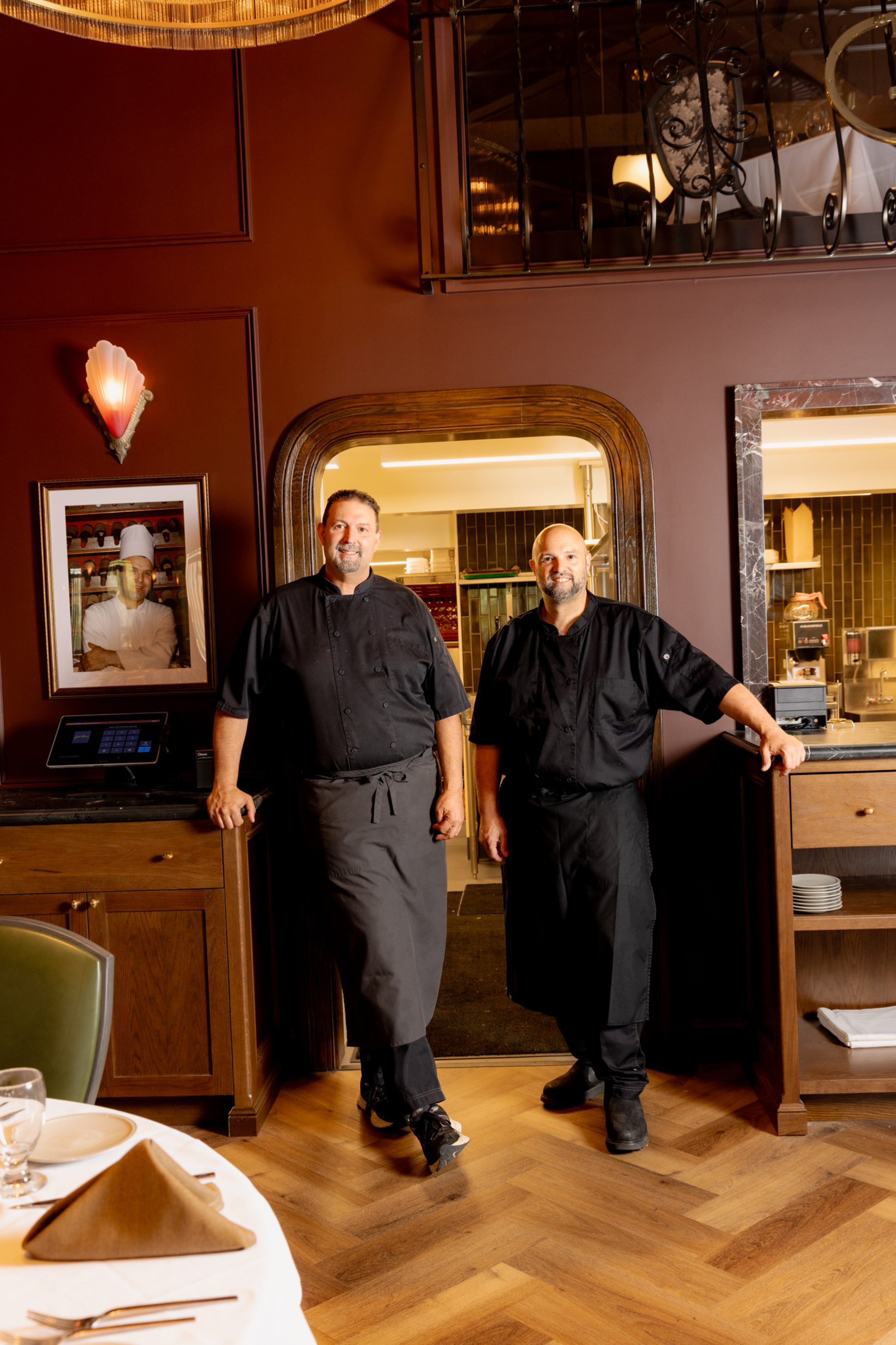 Two chefs in black uniforms stand in a restaurant doorway, surrounded by warm wood tones and elegant dining room decor.