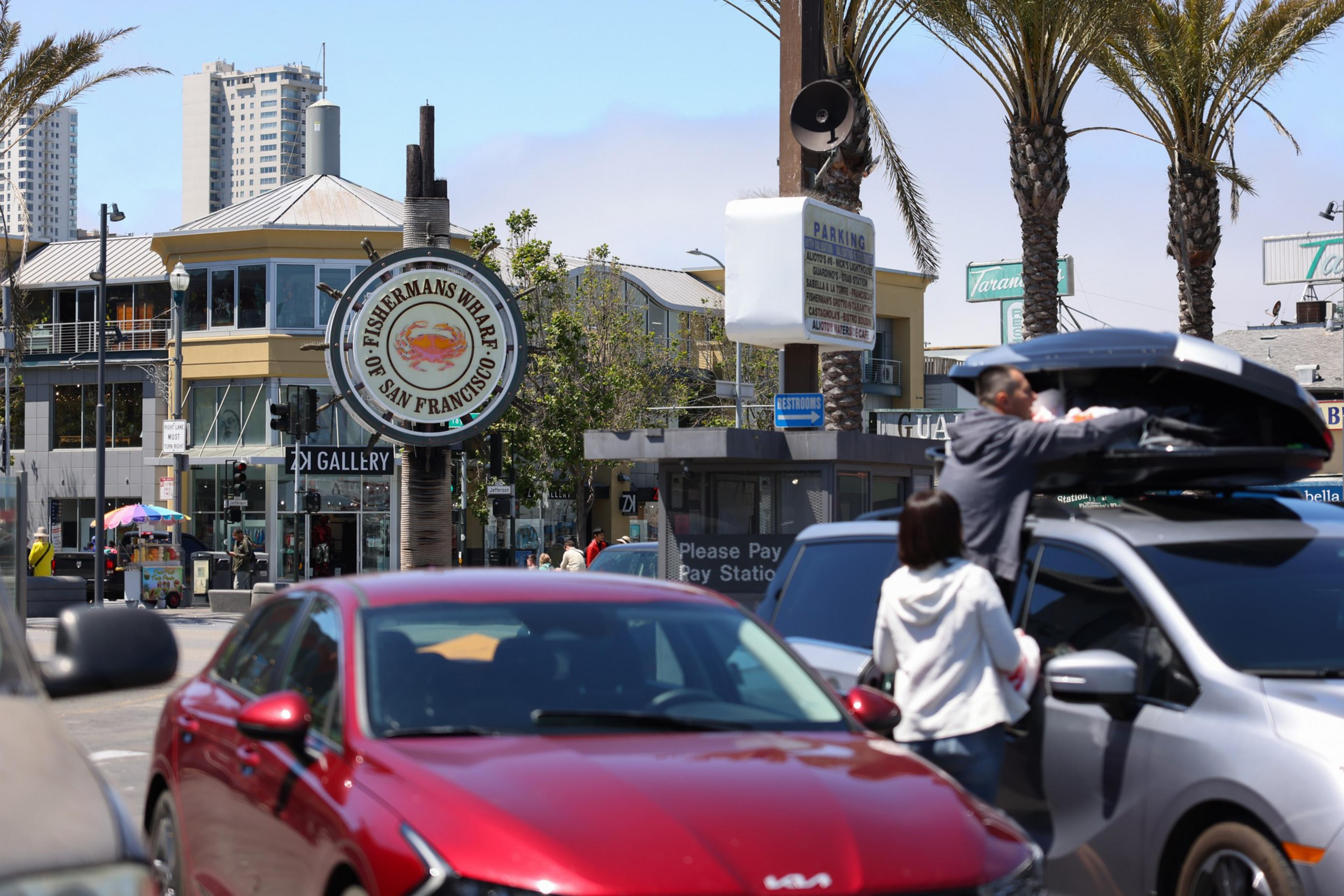 A busy street scene near Fisherman's Wharf in San Francisco, showing cars, palm trees, and people loading a rooftop storage box on a gray vehicle.