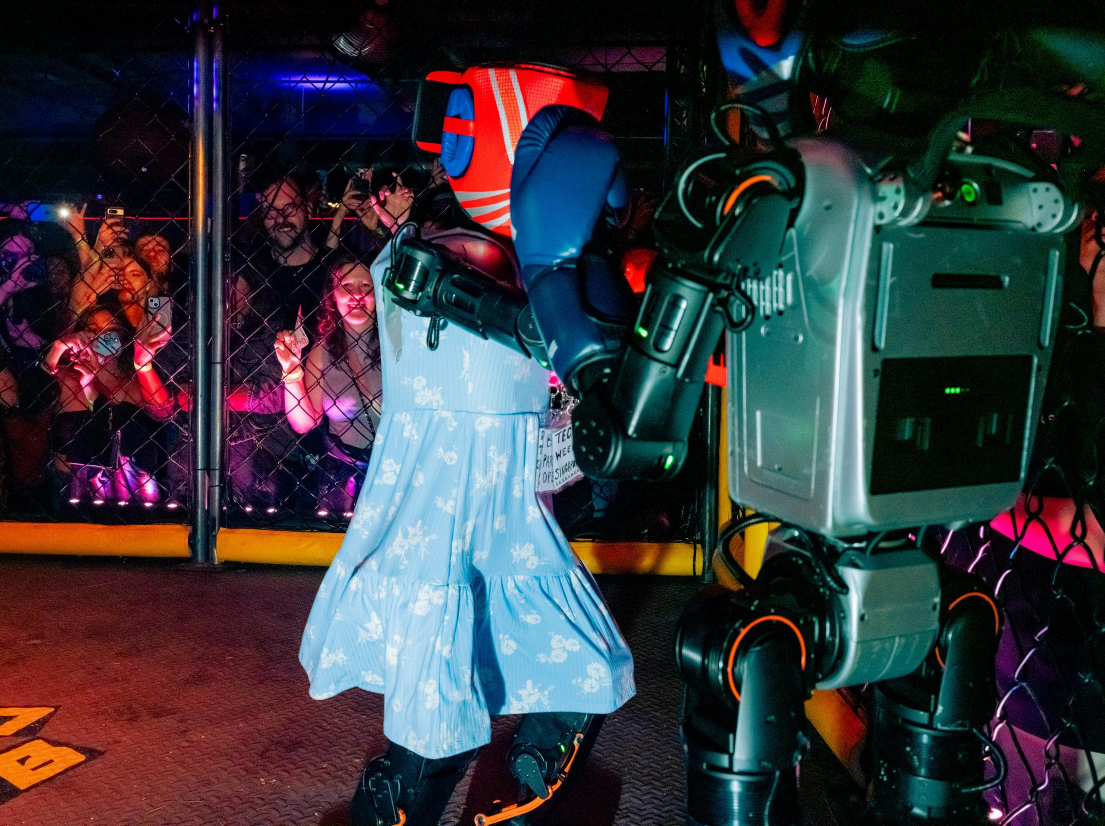A robot wearing a blue floral dress and red boxing gloves fights another metallic robot inside a cage with a crowd watching behind.