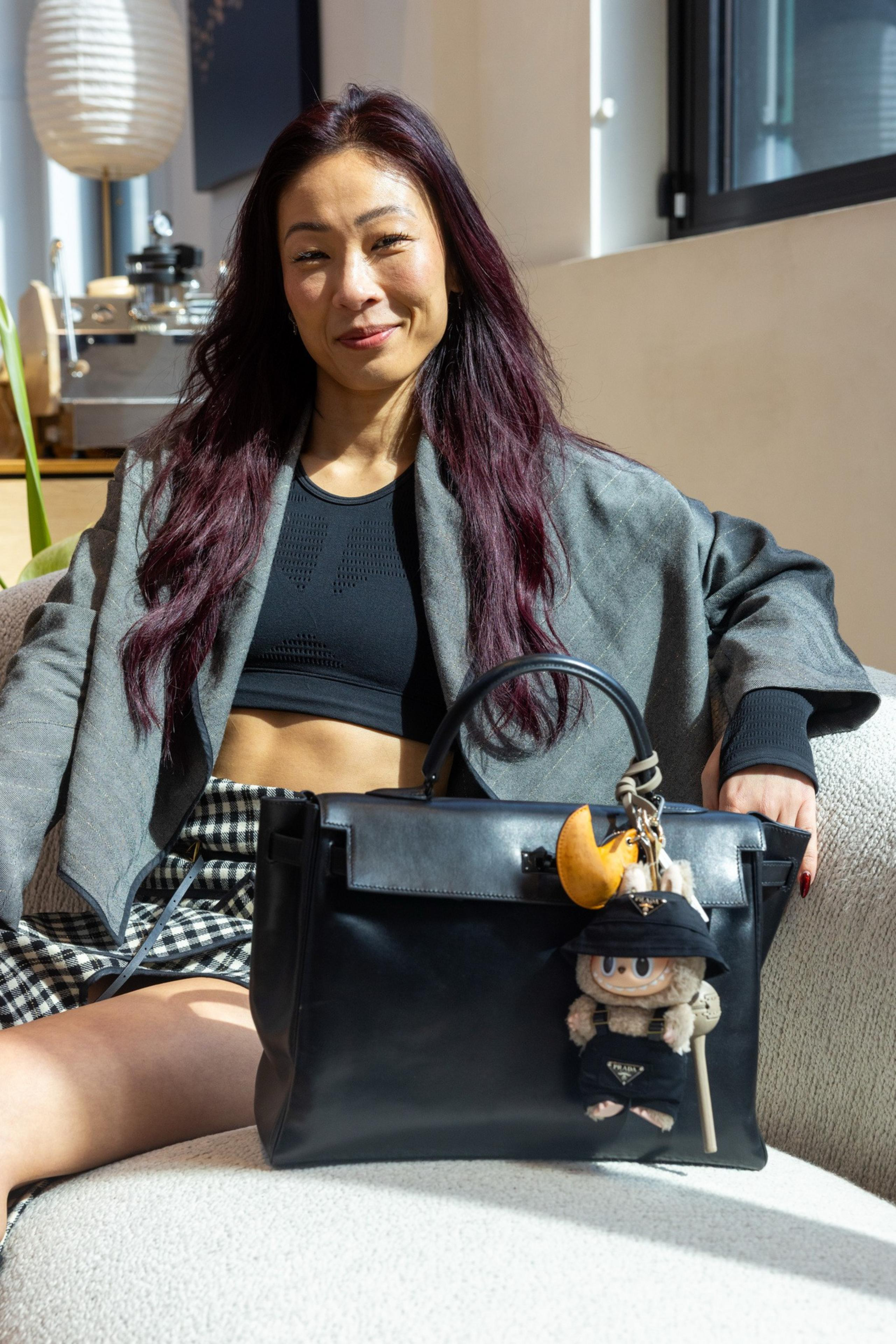 A person with long, dark red hair sits on a chair. They're wearing a black top, gray blazer, and checkered skirt. A black handbag with a doll charm is beside them.
