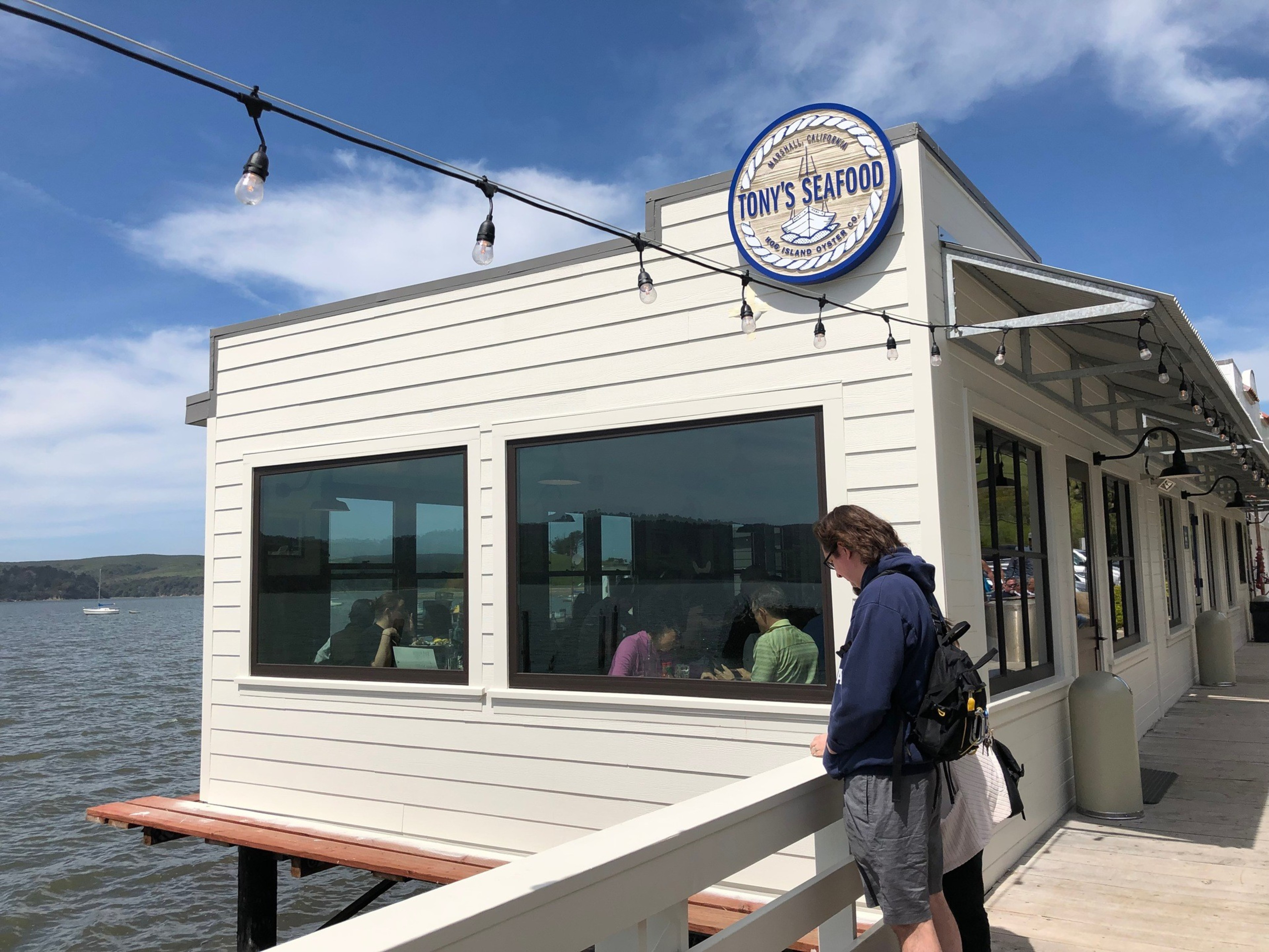 A woman stands next a small restaurant on the water. 
