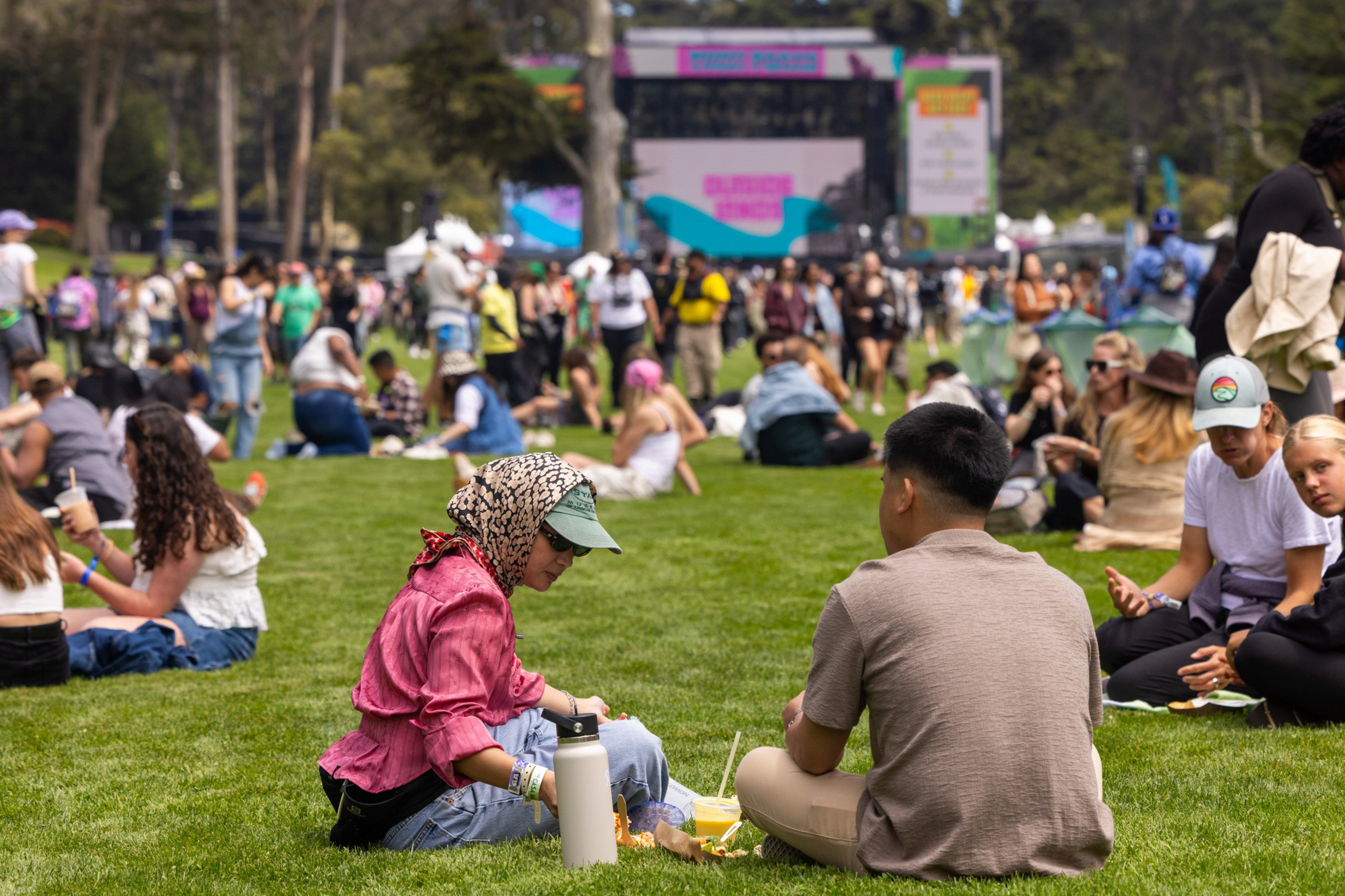 People sit on the grass in a park, eating and socializing, with a large stage and more crowd visible in the background.
