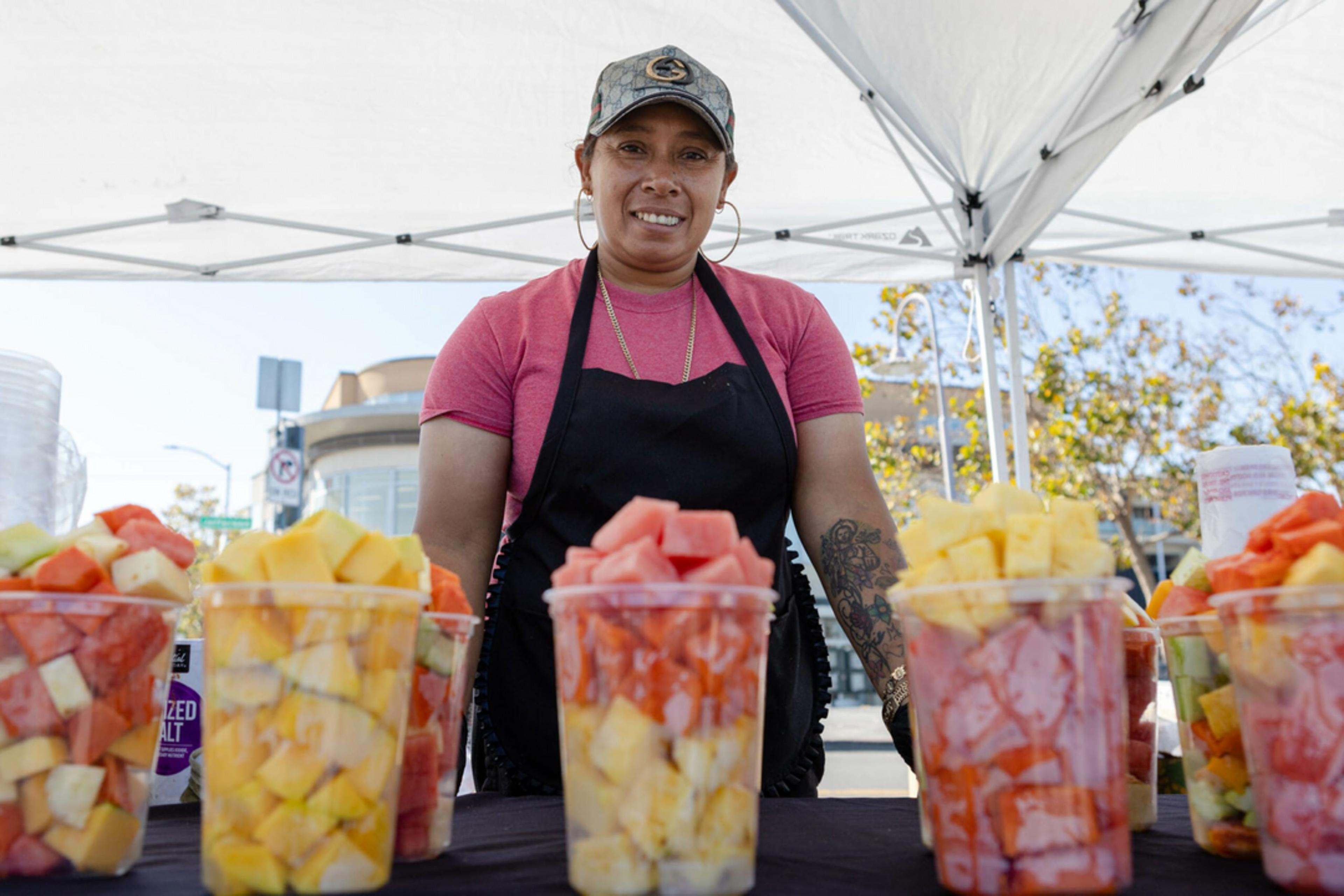 A person wearing a pink shirt, black apron, and camo cap stands behind a table with clear cups filled with mixed fruit under a white canopy.