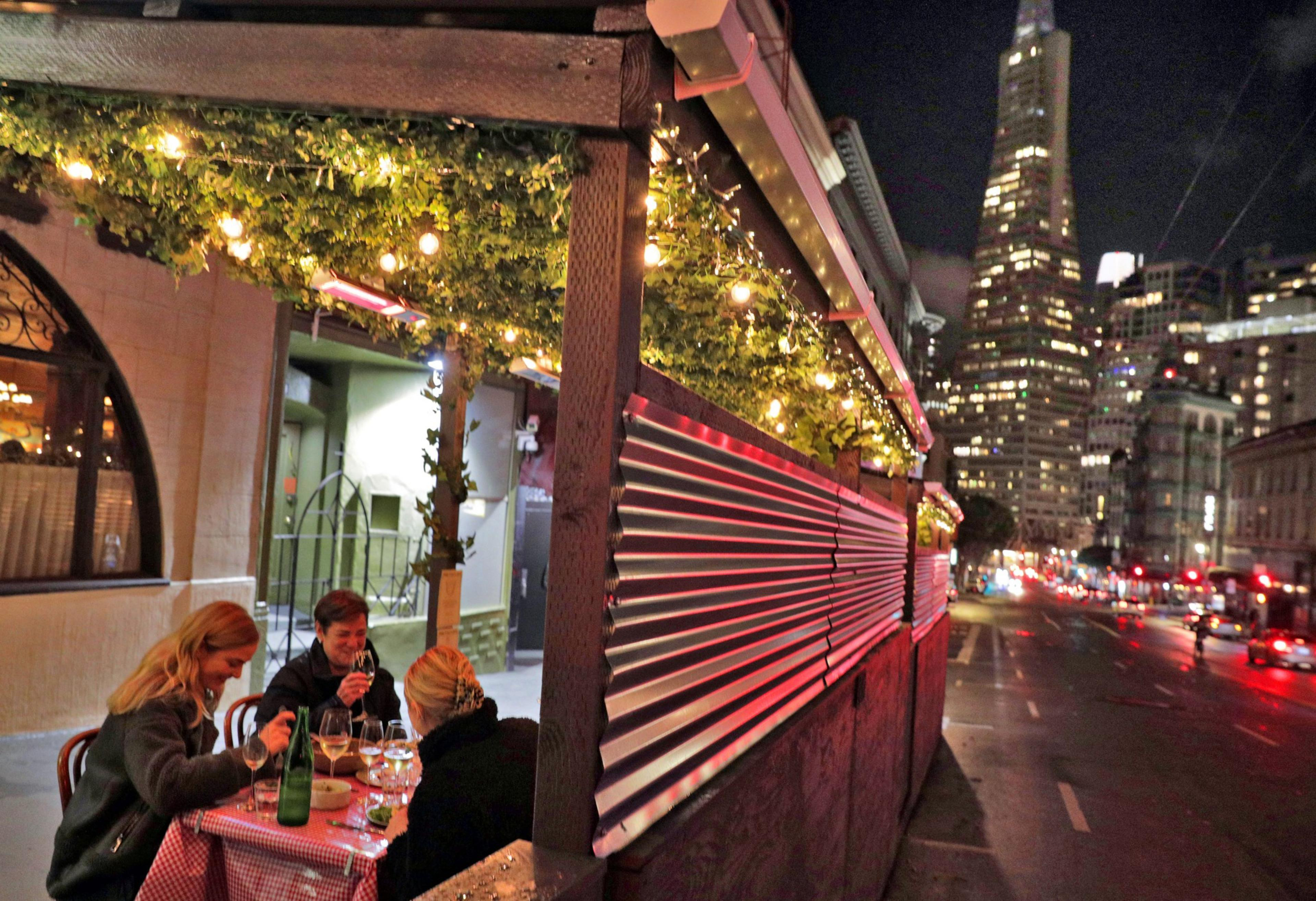 Three people are dining outside under string lights at a table with a checkered cloth. A city street and a tall illuminated building are visible at night.