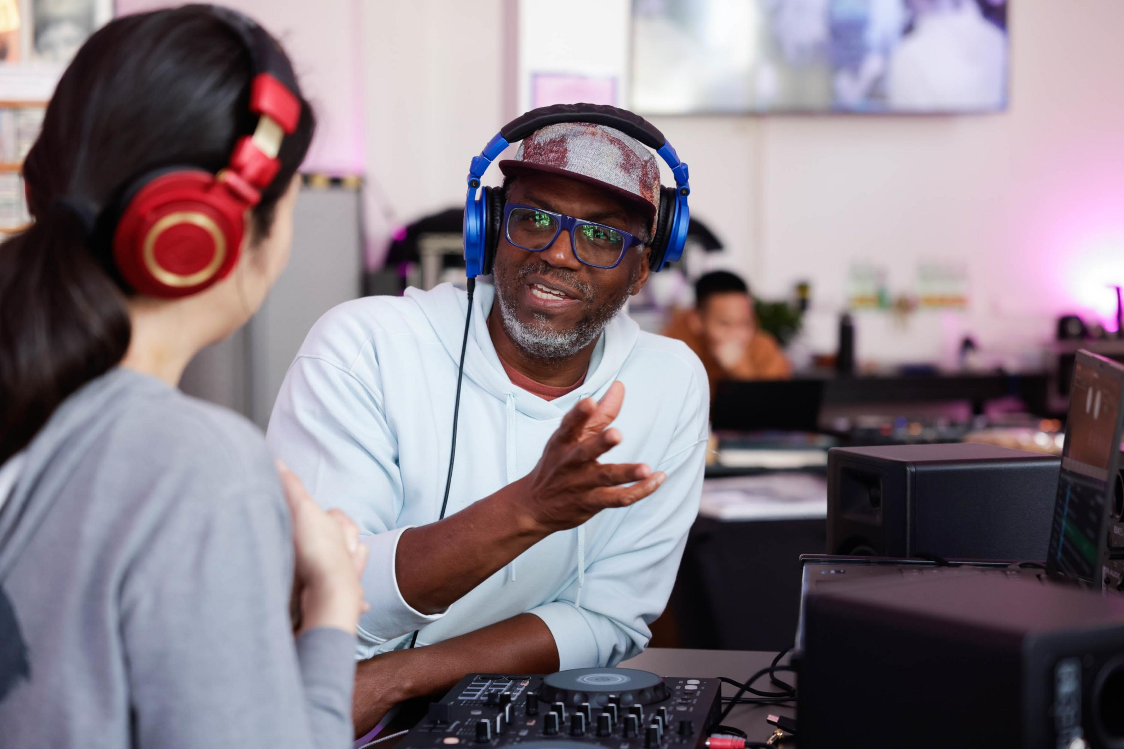 A man wearing headphones speaks to a woman with red headphones in a studio setting with audio equipment.