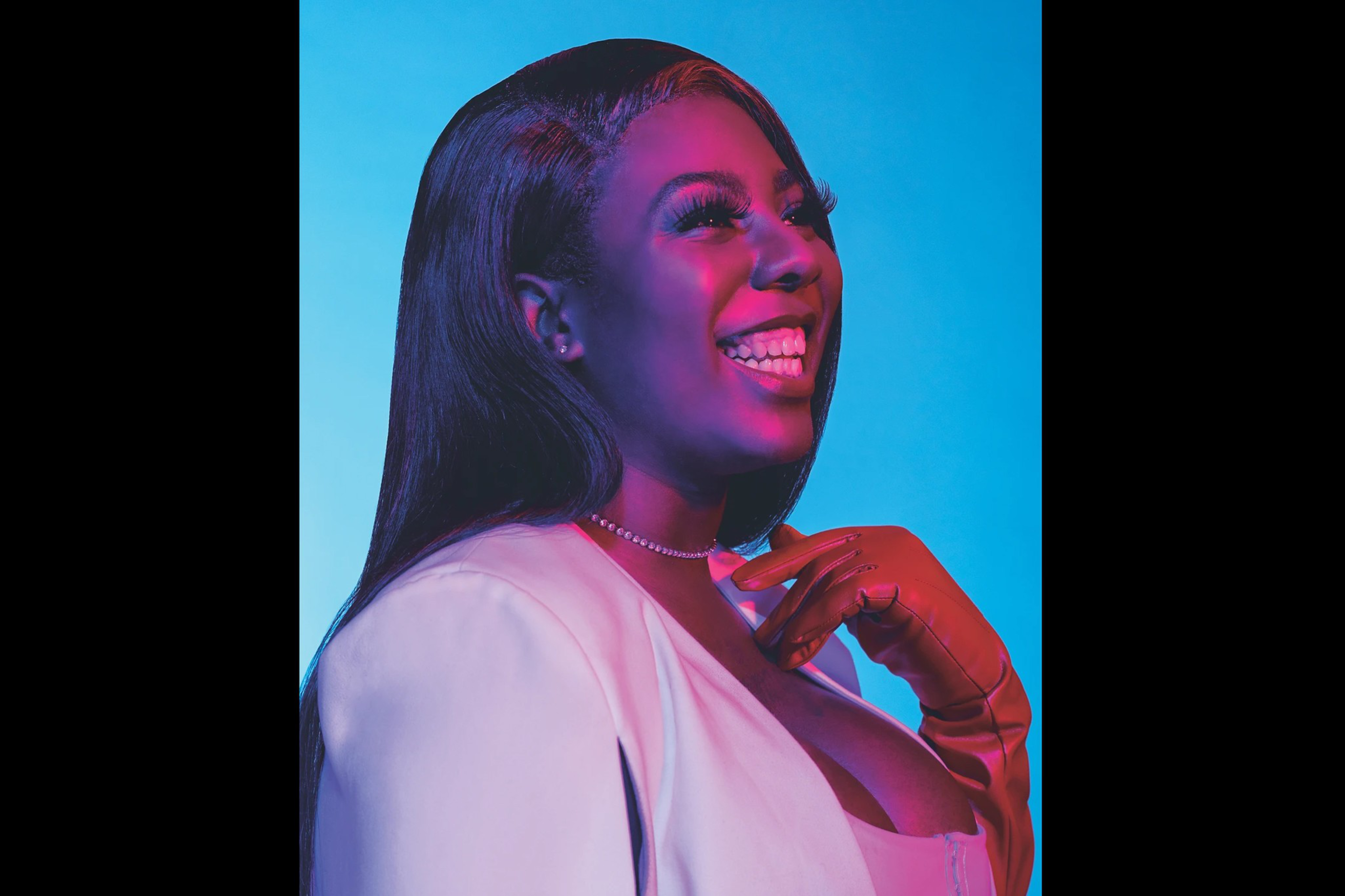 A woman with long straight hair smiles brightly, wearing a light-colored blazer, a necklace, and her hand near her neck against a blue background.