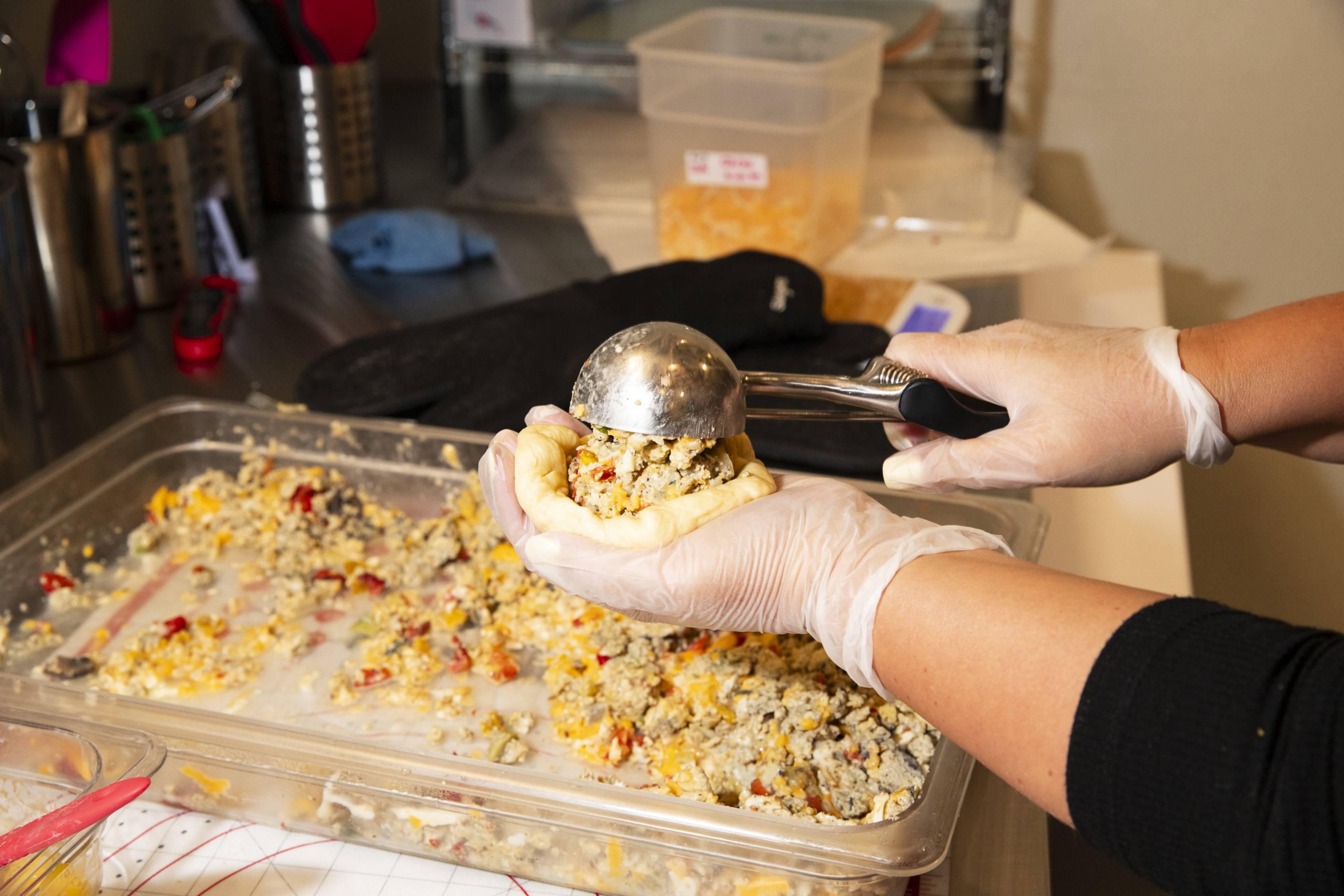 Gloved hands hold a dough cup while scooping a colorful mixture of meat, cheese, and vegetables from a large clear container in a kitchen setting.