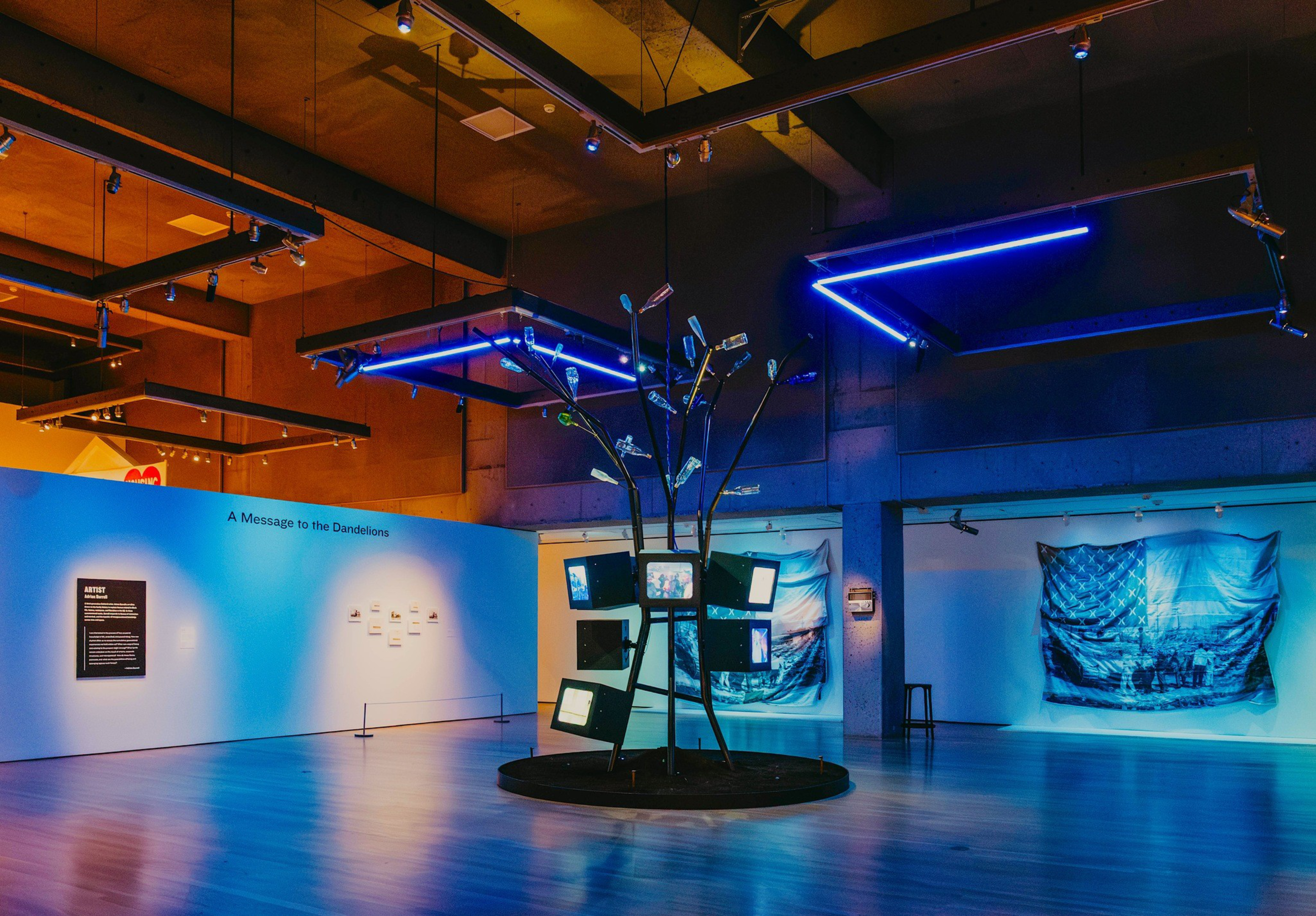 Multiple TV screens are mounted on a leafless metal tree sculpture under blue neon lights in a spacious gallery with blue-lit art pieces on the walls.