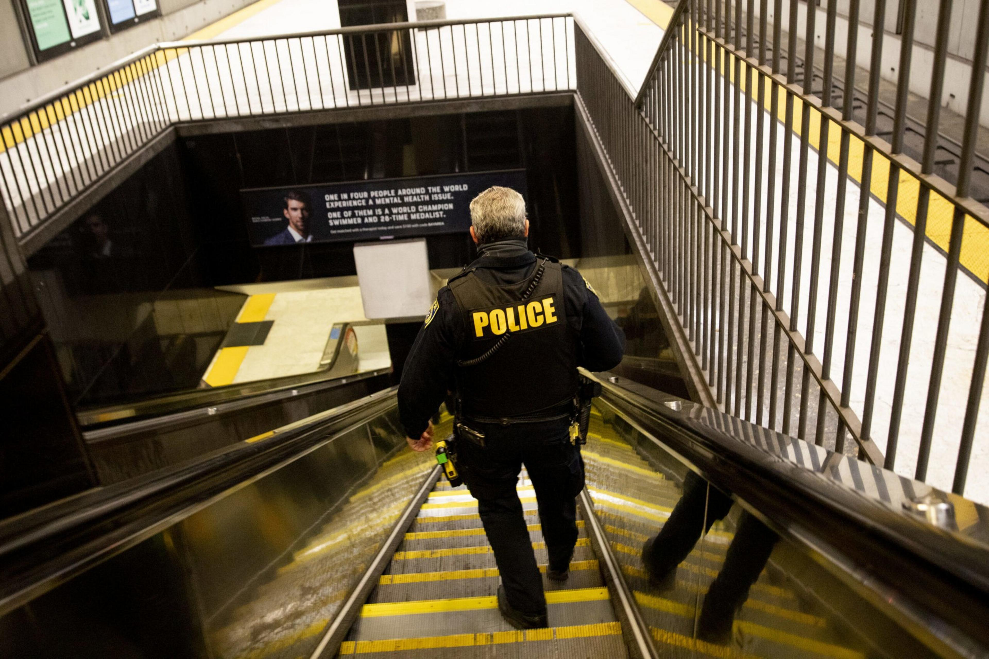 A police officer in uniform descends an escalator in a subway station, with metal railings on both sides and a mental health awareness poster ahead.