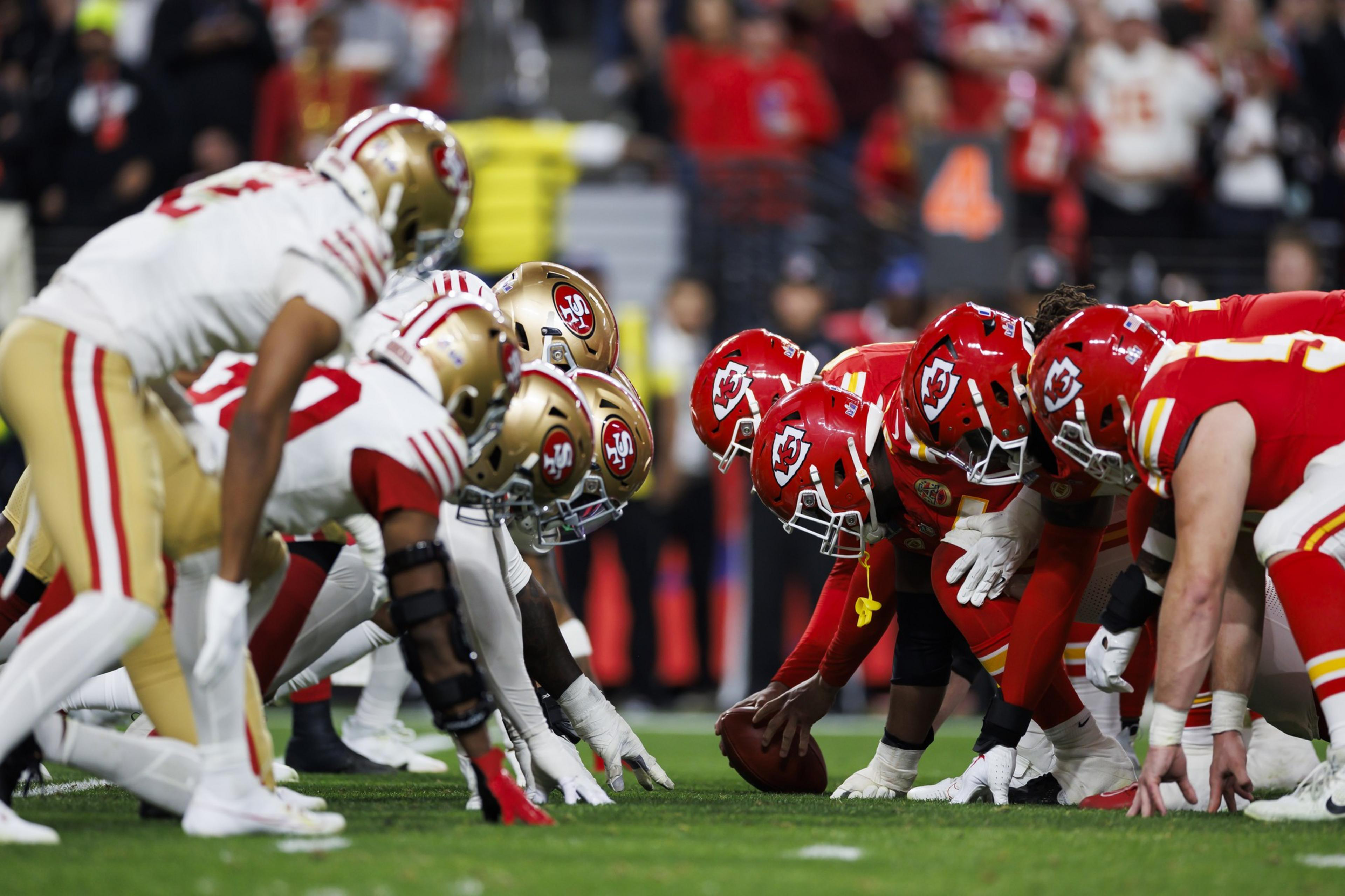Two American football teams face off at the line of scrimmage, wearing red and gold uniforms. Players are crouched, focusing on the snap, with an audience in the background.