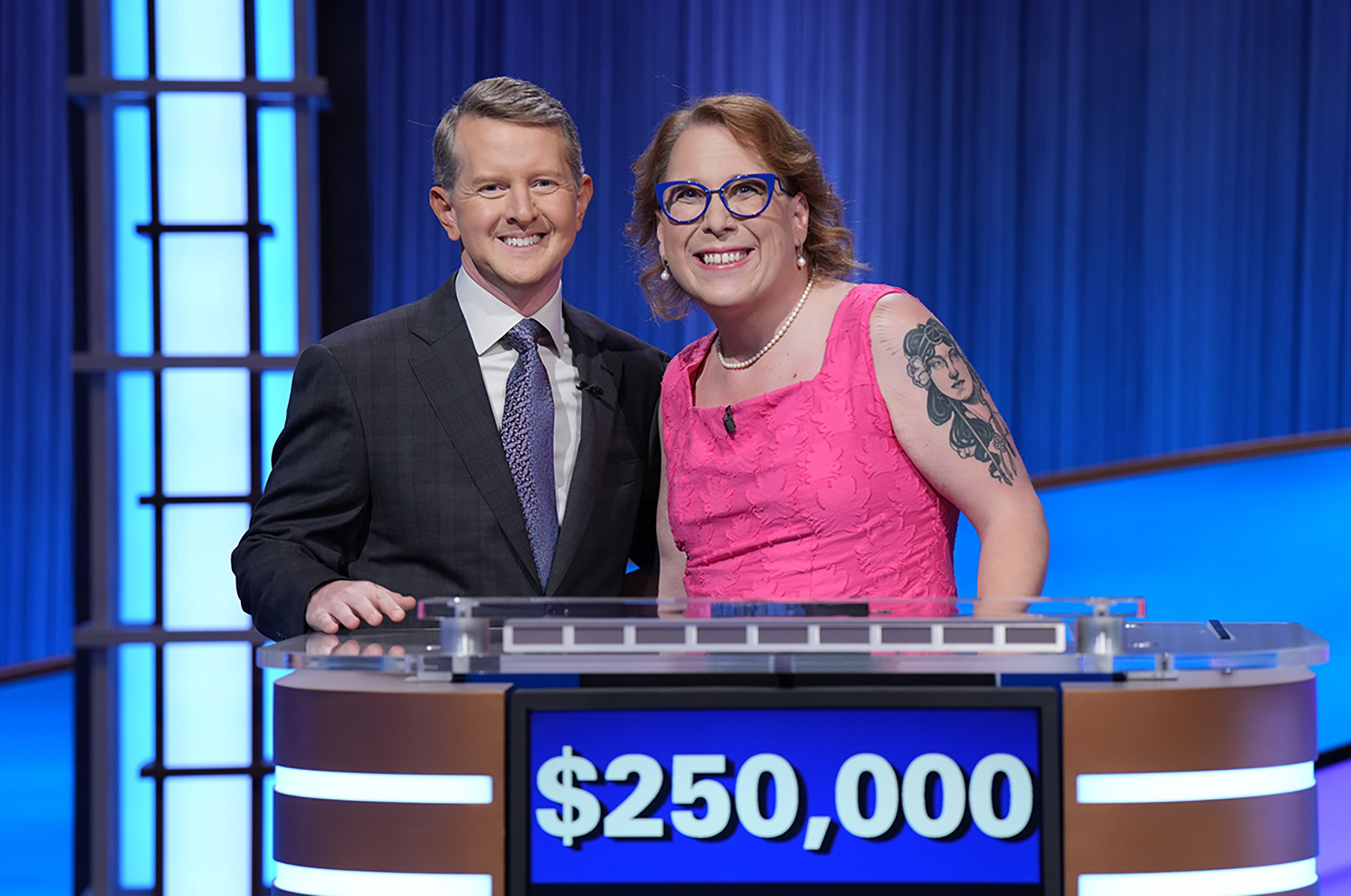 A man in a suit and a woman in a pink dress with glasses and a tattoo smile behind a game show podium displaying $250,000.