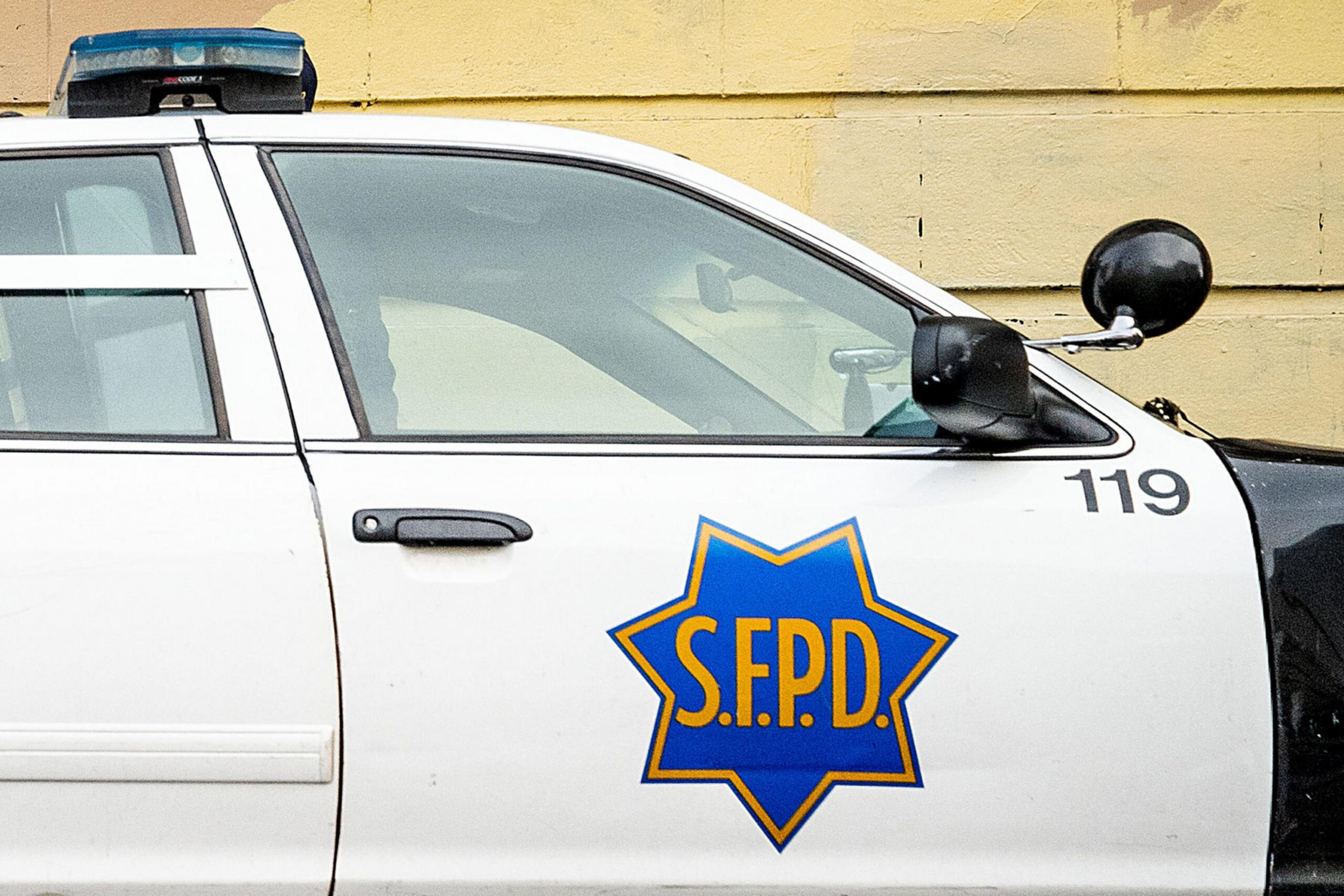 A white police car with the blue and gold emblem of "SFPD" on the side, marked with the number 119, parked against a yellow wall.
