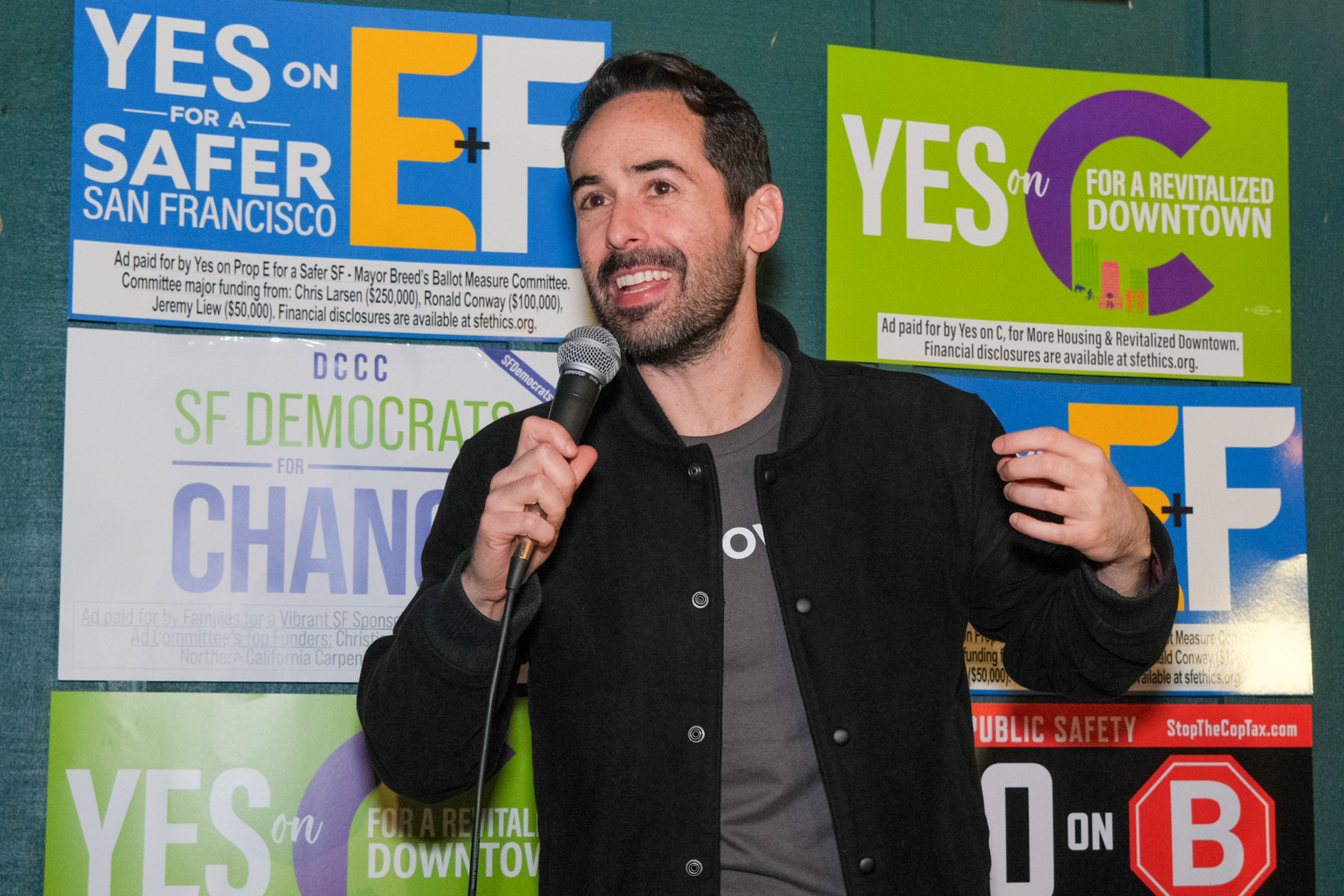 Grow SF co-founder Steven Buss holds a microphone and smiles in front of campaign signs.