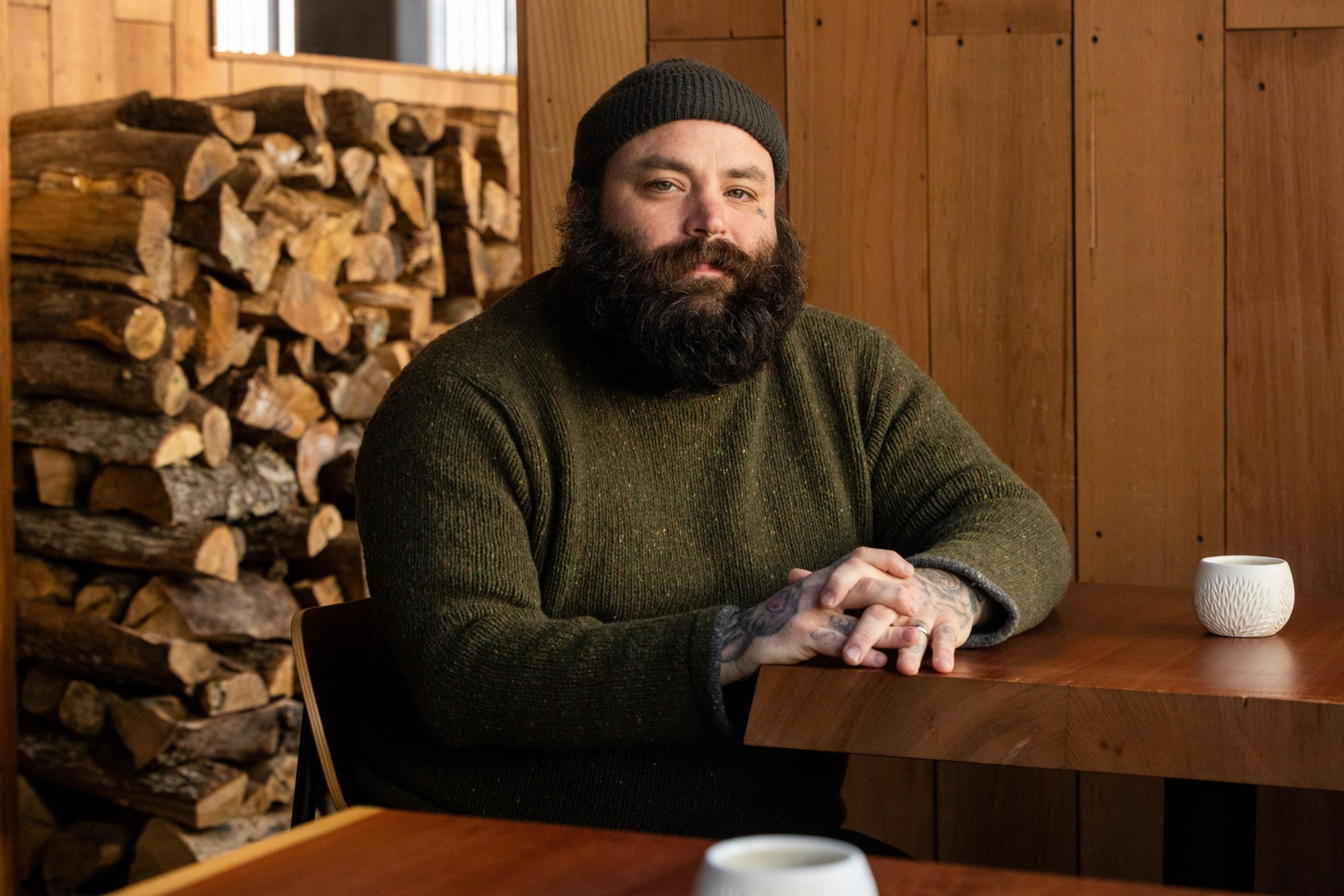 A bearded person in a green sweater and beanie sits at a wooden table, hands clasped. A pile of firewood is stacked behind them, with a white mug on the table.