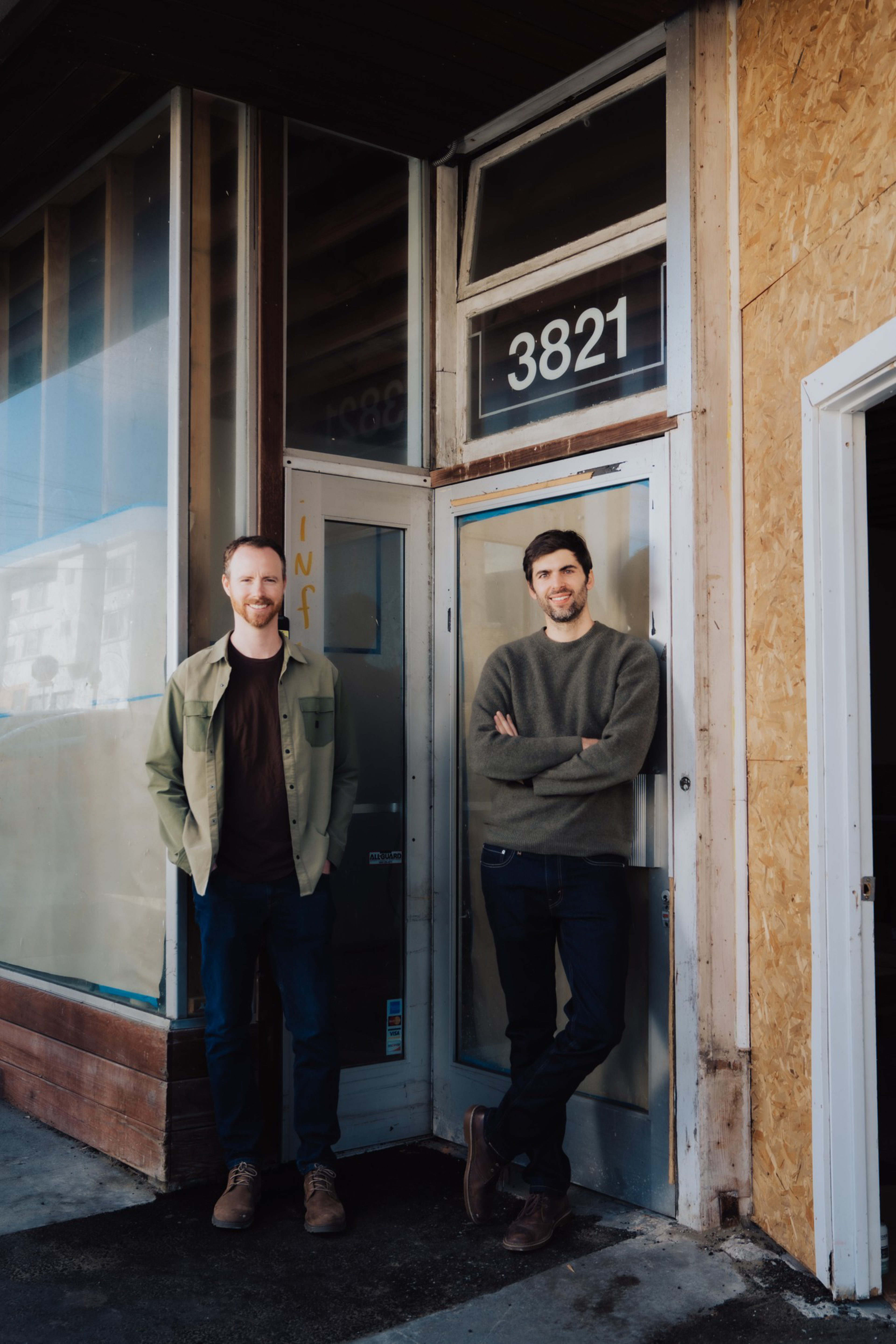Two men stand outside a storefront numbered 3821, one with hands in pockets wearing a green jacket, the other leaning with arms crossed in a gray sweater.