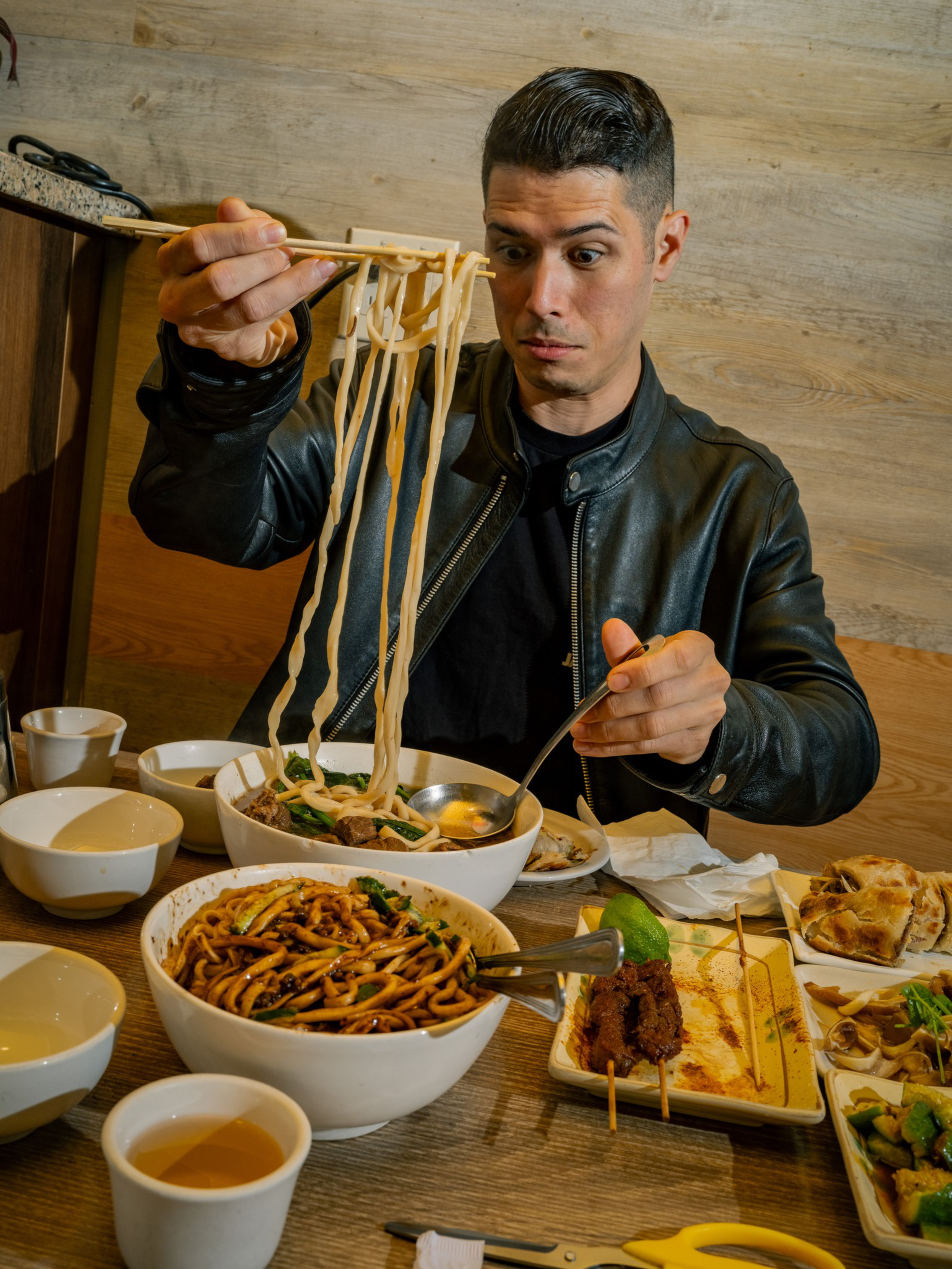A man in a black leather jacket lifts long noodles with chopsticks from a bowl, surrounded by various dishes and bowls on a wooden table.
