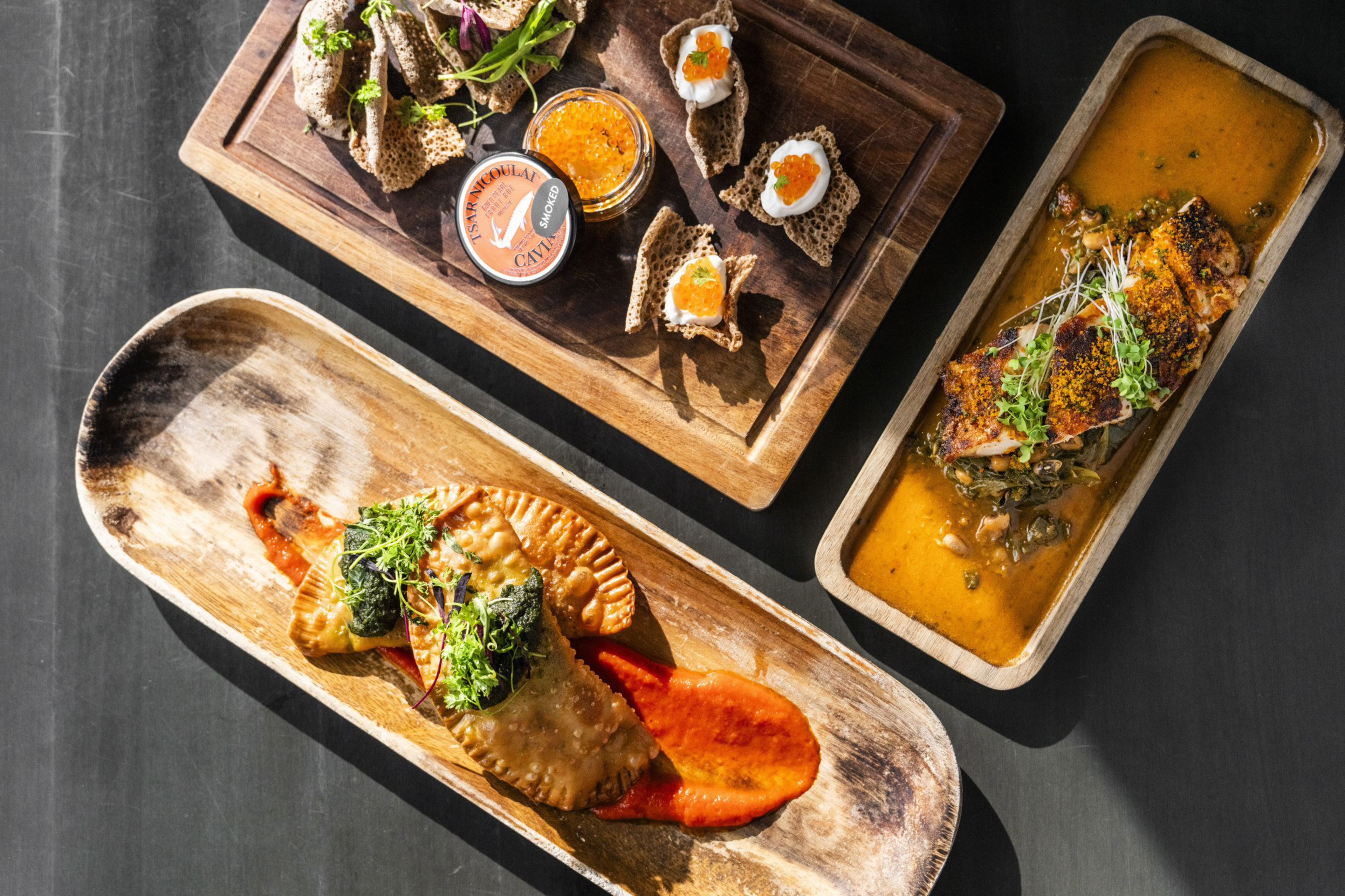 The image shows three wooden serving trays with various dishes: empanadas on red sauce, salmon with herbs in broth, and buckwheat blinis with caviar and cream.