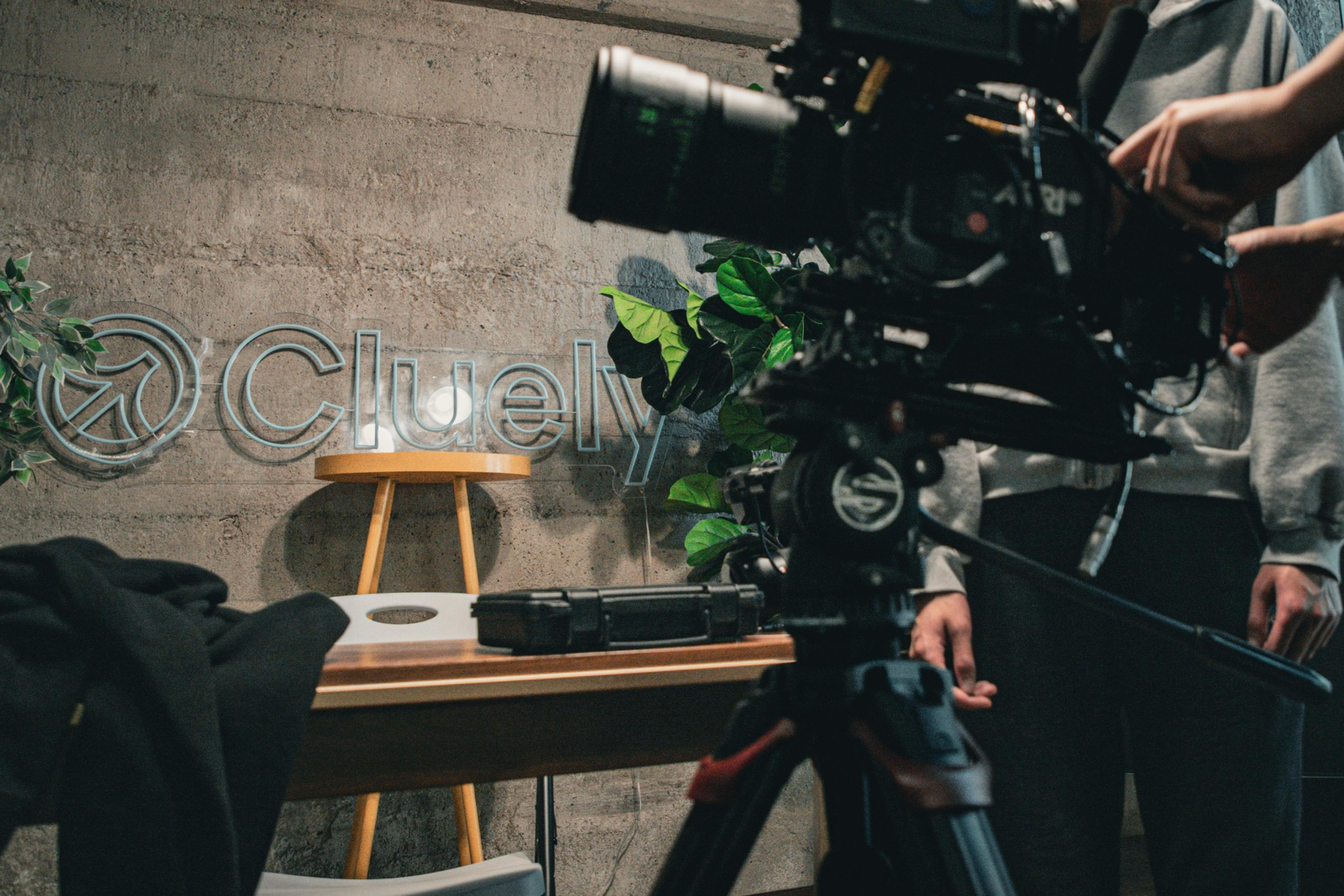 A professional camera setup is arranged in front of a table with a suitcase, chair, and a wall logo reading &quot;Cluely&quot; against a concrete background with plants.