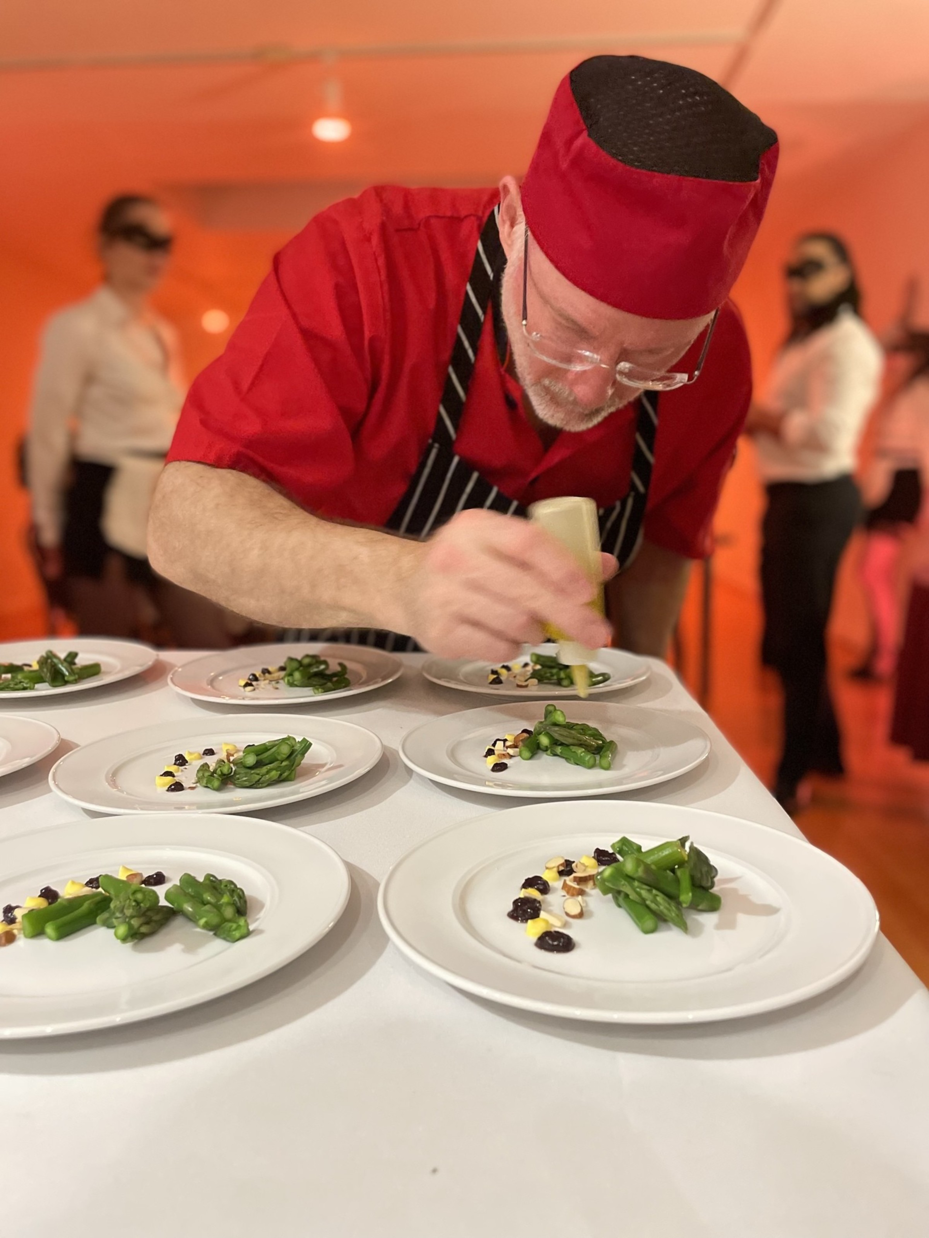 A chef in a red uniform and hat carefully garnishes plates with asparagus and small dollops of sauce in a warmly lit kitchen.