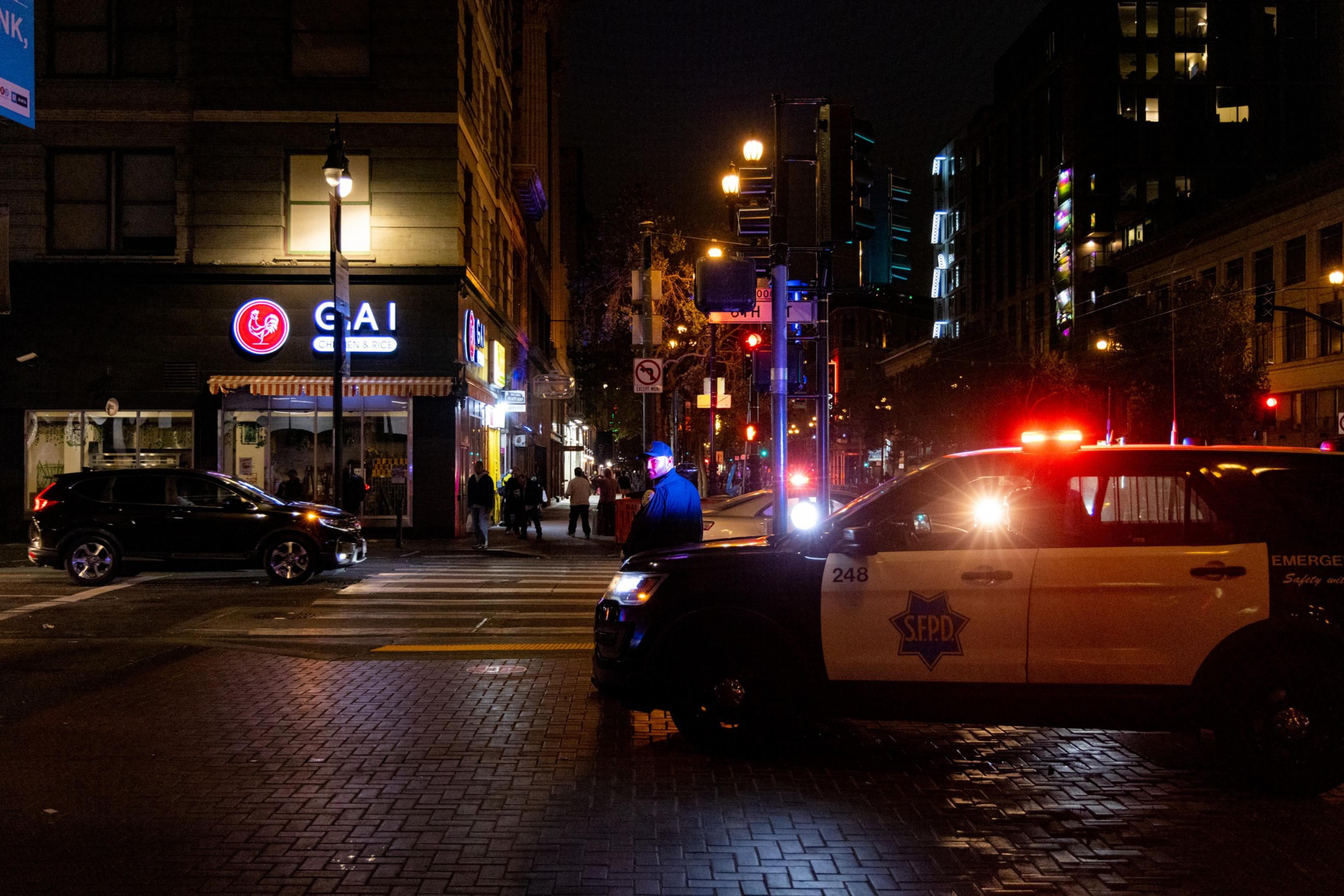 A nighttime street scene shows a police car with flashing lights, an officer standing by, a lit "GAI Chicken & Rice" sign, and people walking on the sidewalk.