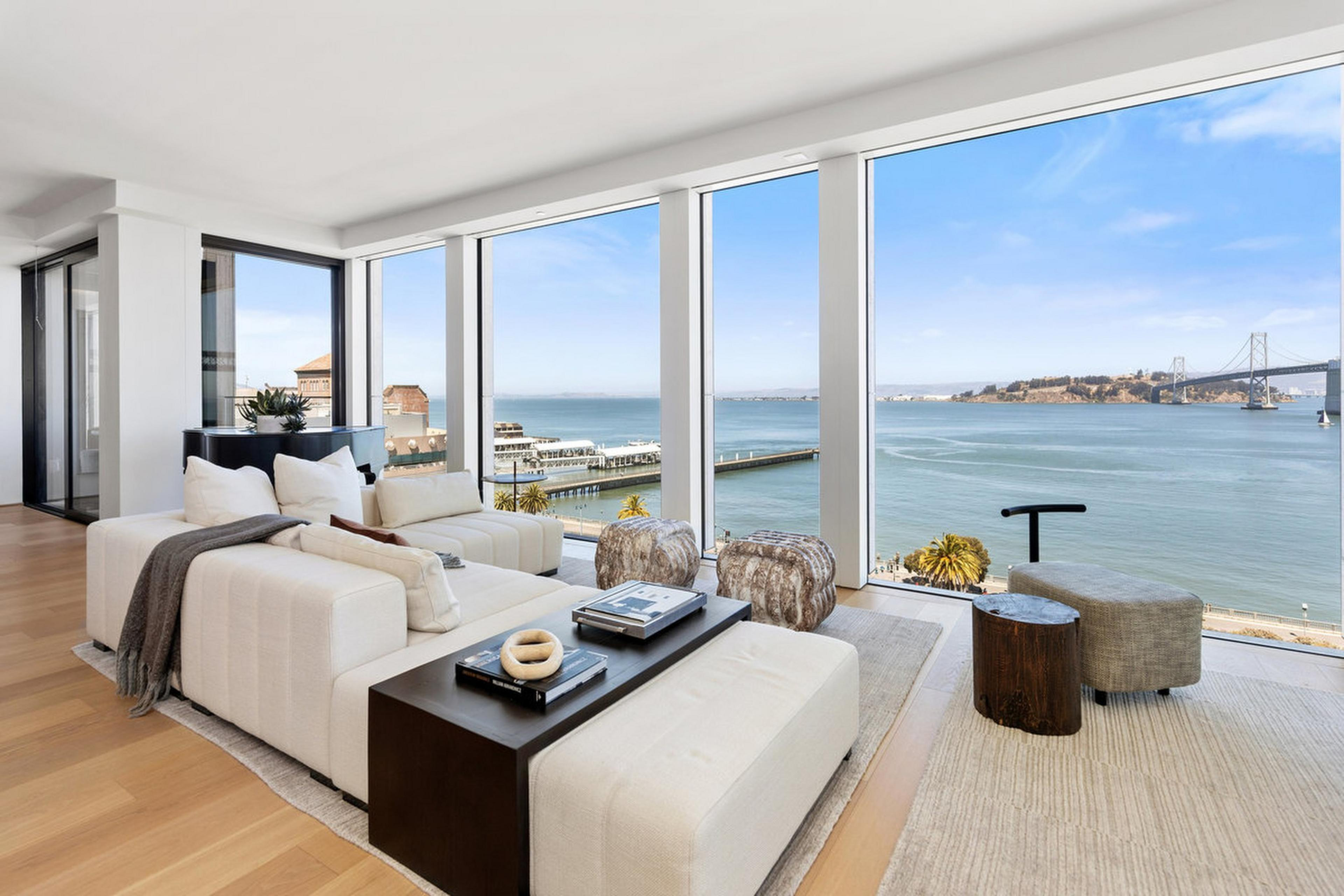 A modern living room with white sofas, a dark wood coffee table, and large floor-to-ceiling windows offering a view of the ocean, pier, and a distant bridge.
