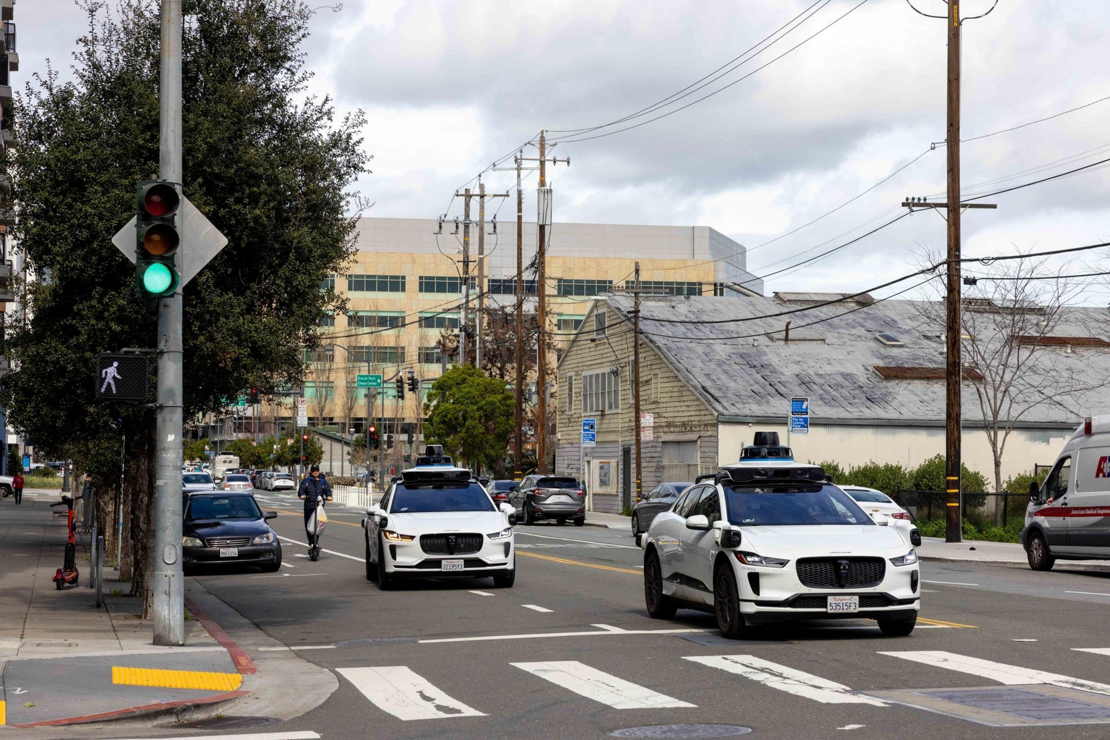 Two self-driving cars and a person on a scooter move on a city street. A green traffic light and a pedestrian crossing are visible. Buildings and trees line the road.
