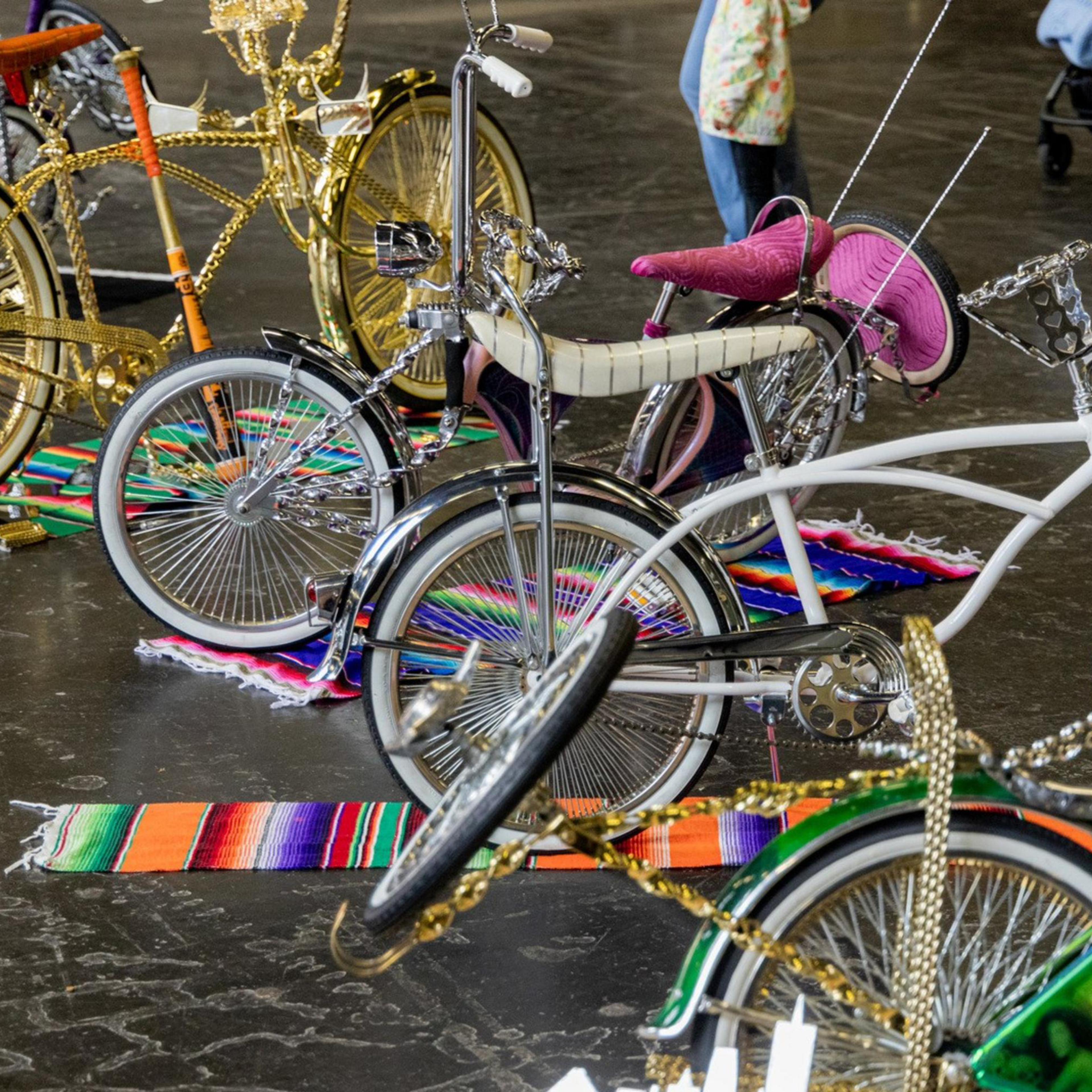 The image shows a display of customized lowrider bicycles with elaborate designs and colorful accessories, set on vibrant blankets in an indoor exhibition space.
