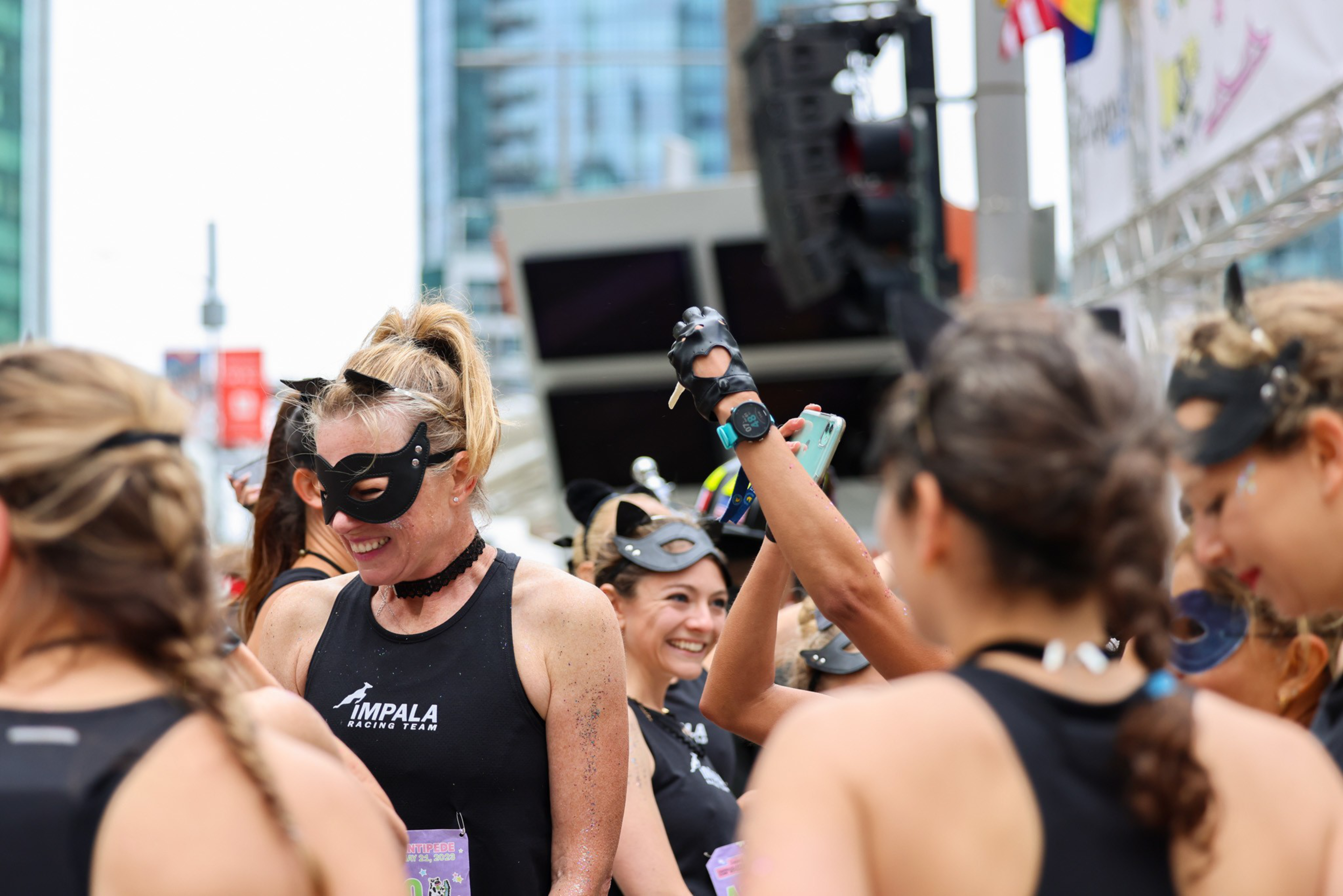 Women dressed in black tank tops and cat masks smile and gather outdoors in an urban setting, engaging in a lively, festive event.