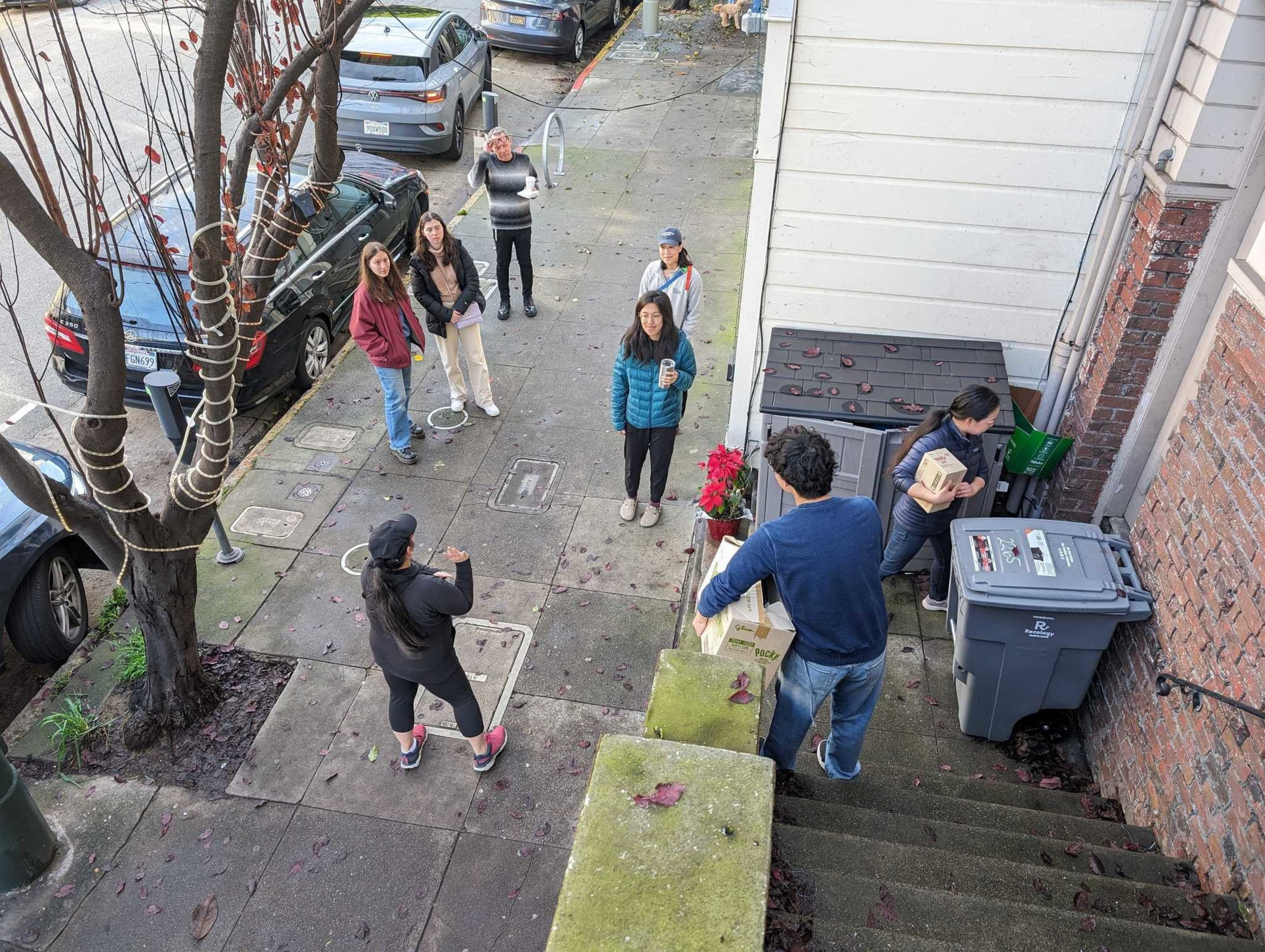 Half a dozen casually dressed people stand on a sidewalk as a man brings a cardboard box full of Pocky biscuit sticks down to distribute.