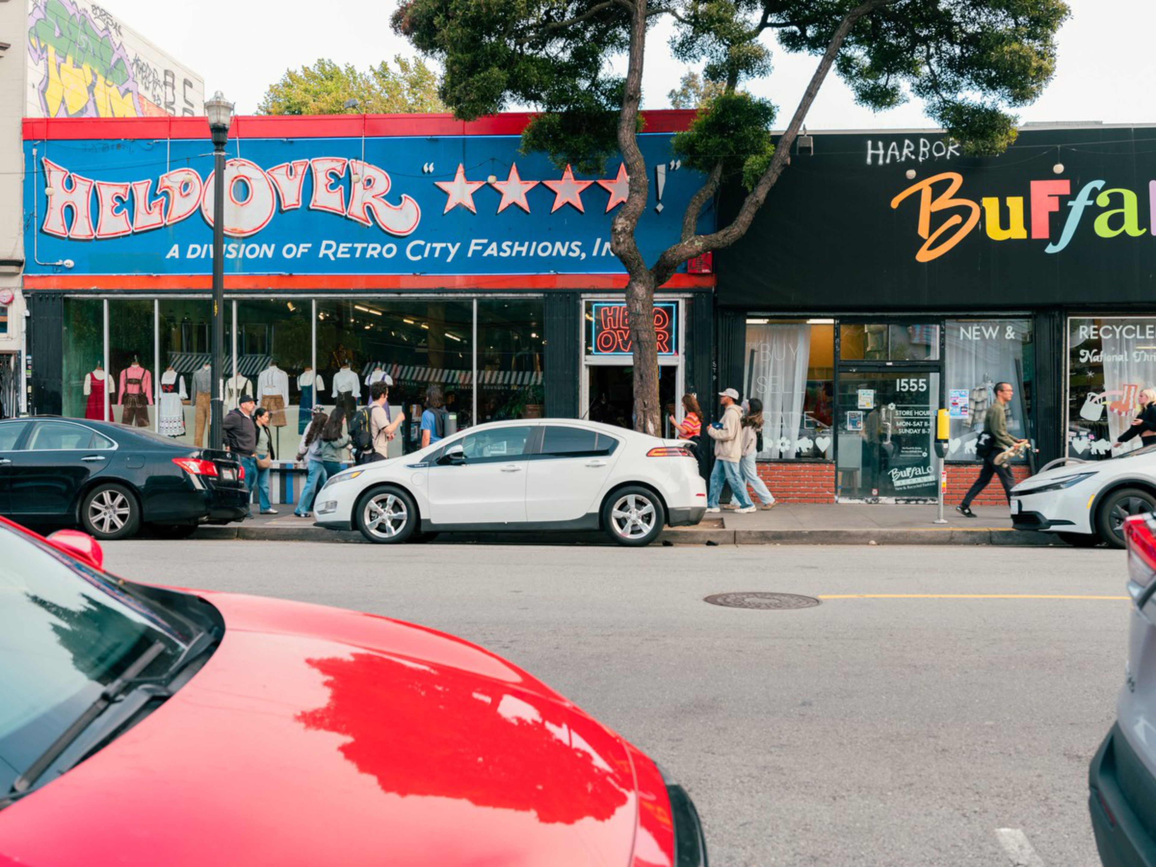 People walk past parked cars in front of two shops, one named "Held Over" with a blue storefront and the other "Harbor Buffalo" with a black storefront.
