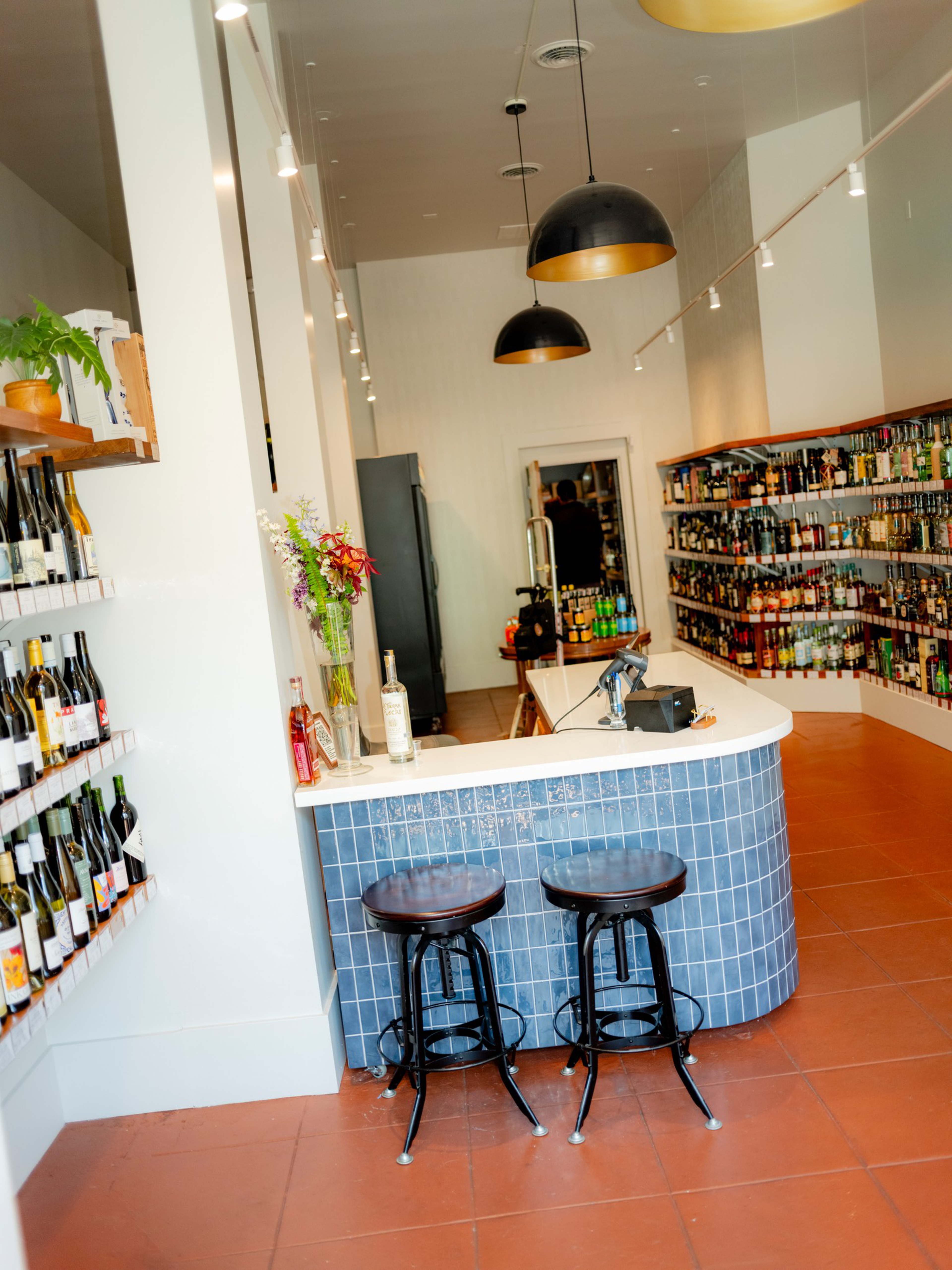 A modern liquor store interior features shelves filled with bottles, a white-topped blue-tiled counter with two stools, pendant lights overhead, and a small vase with flowers.