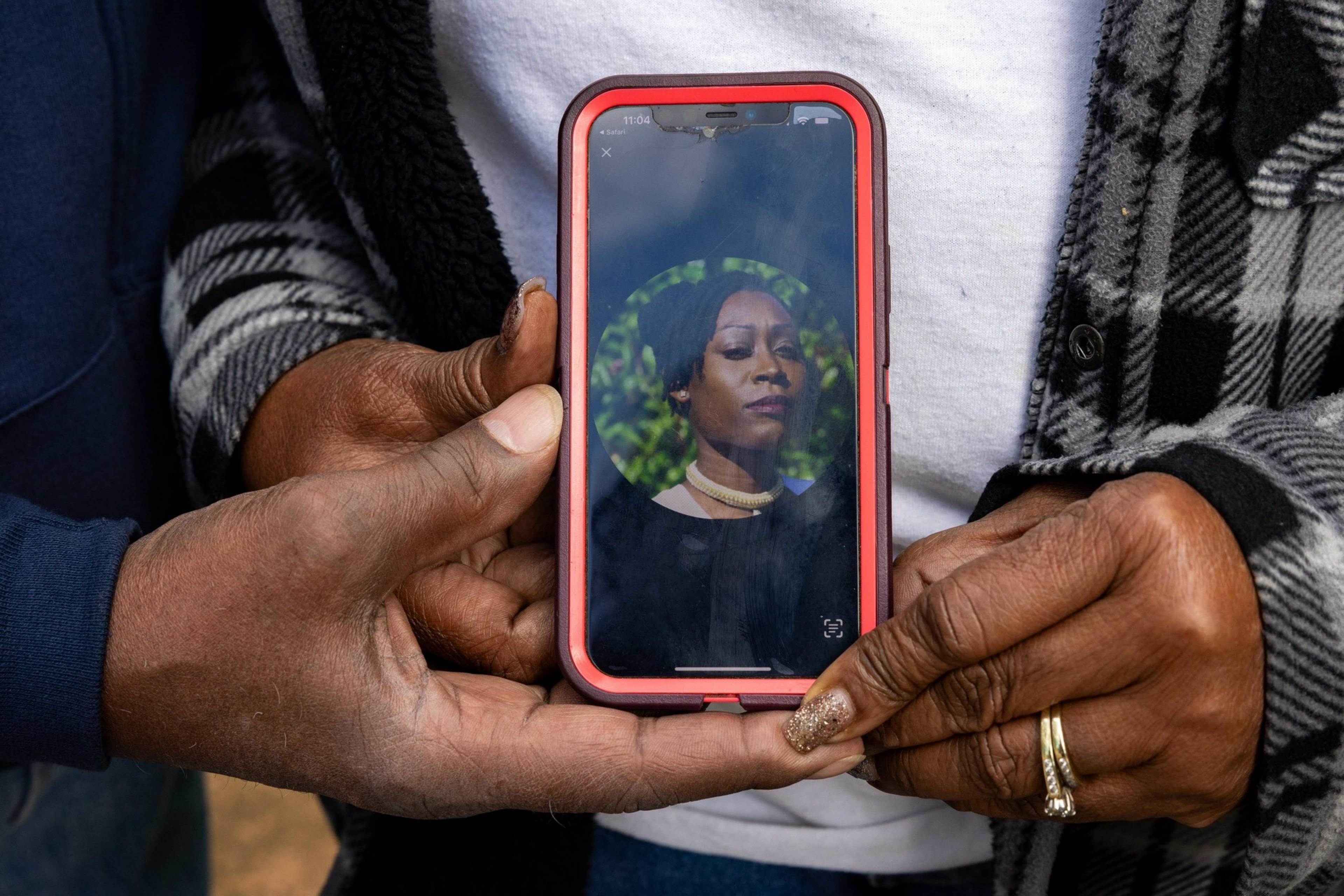 Two hands hold a phone displaying a woman's portrait with a serious expression, set against a green leafy background.