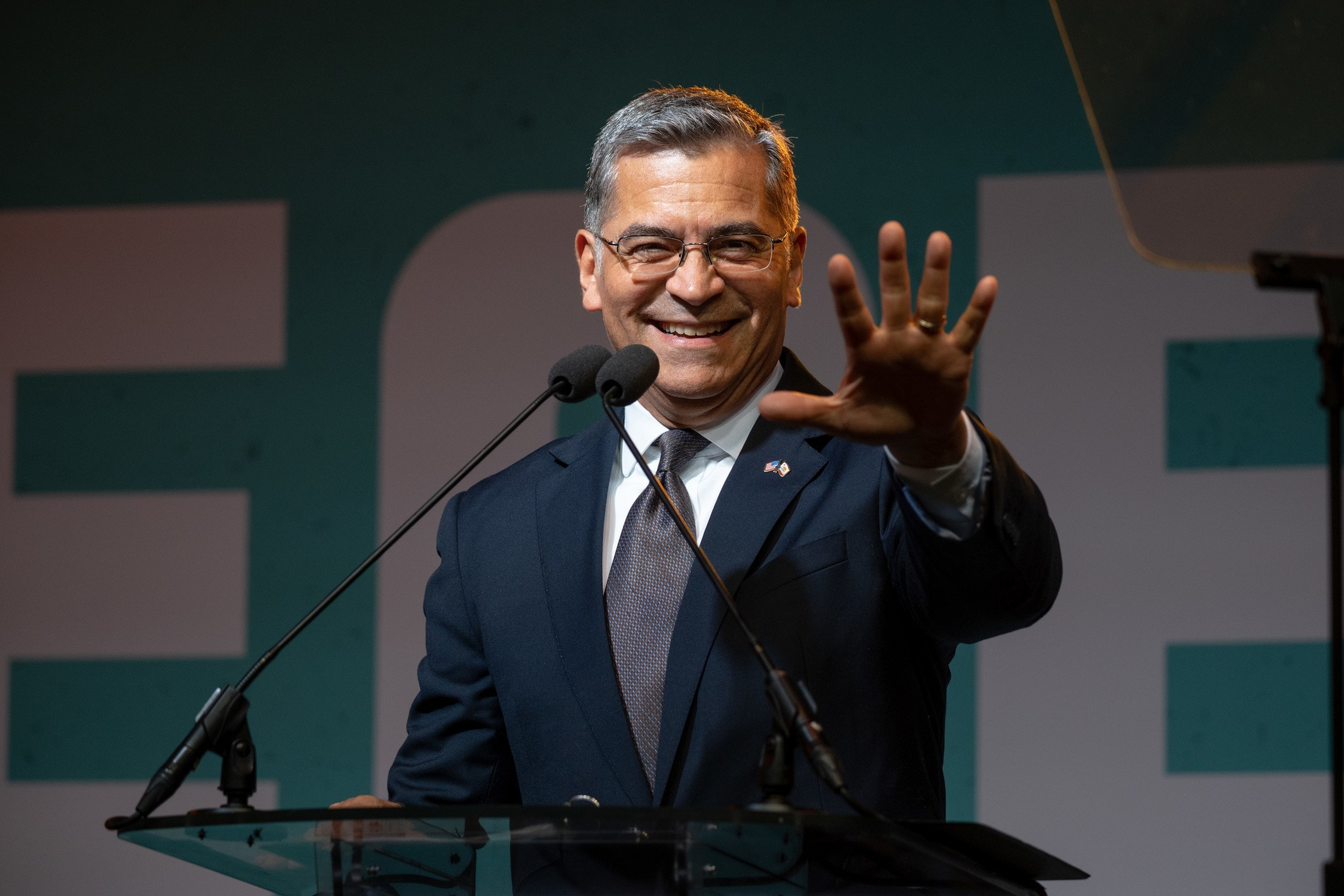 A man in a dark suit and glasses smiles while raising his right hand in front of two microphones at a clear podium.
