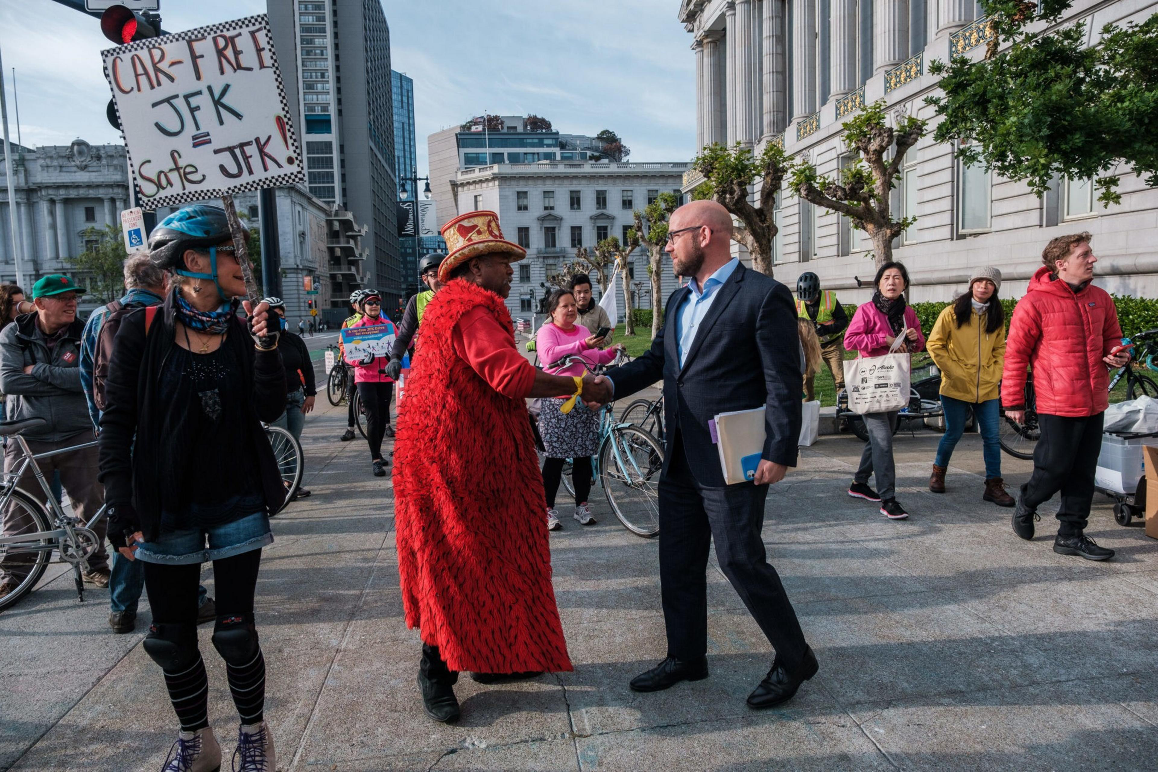 A man in a suit shakes hands with a person in a red coat and hat while people around them hold bikes and a sign reading "CAR-FREE JFK = Safe JFK."