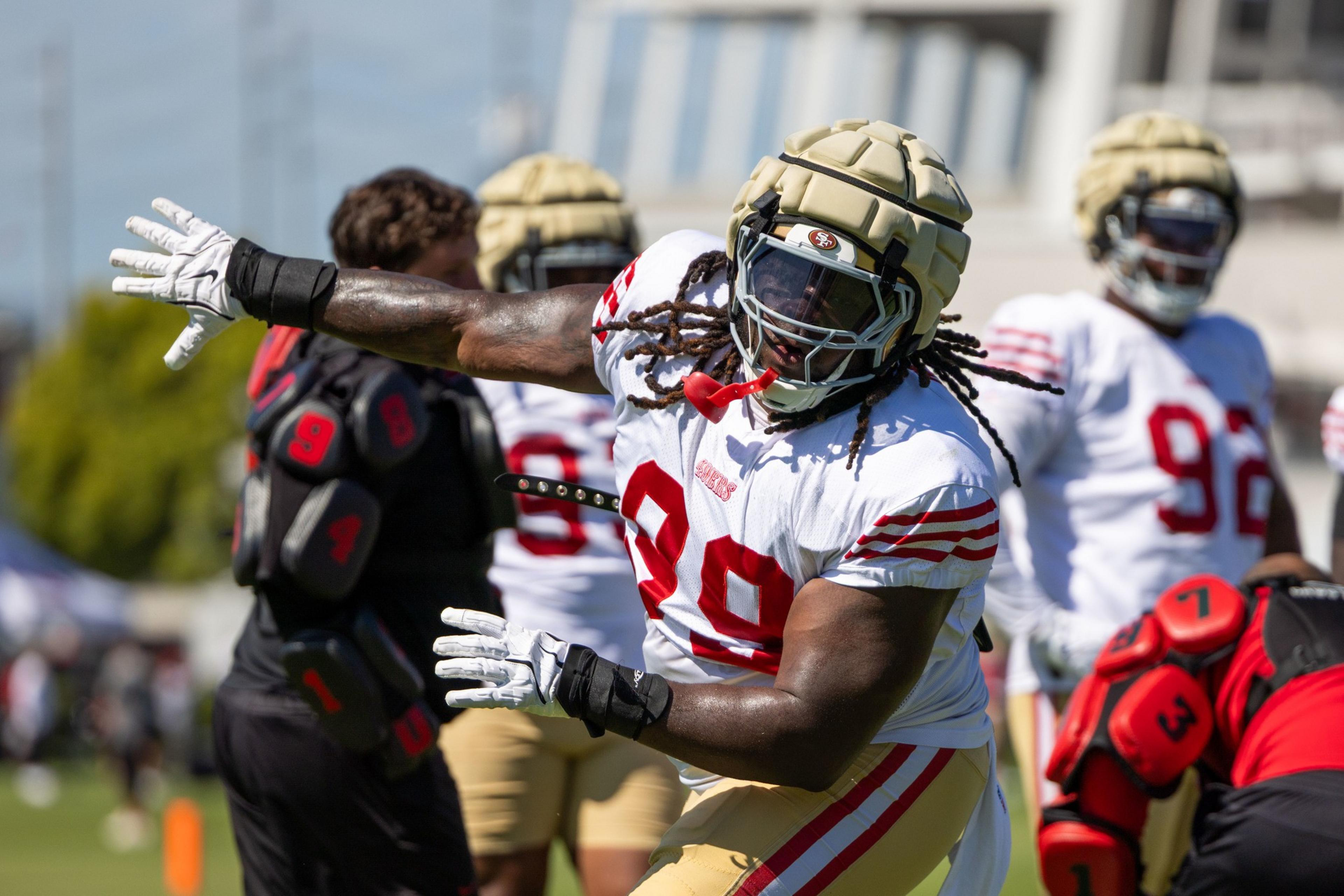 A football player in white and red gear practices on the field. He wears a protective helmet cover and glove, with other players blurred in the background.