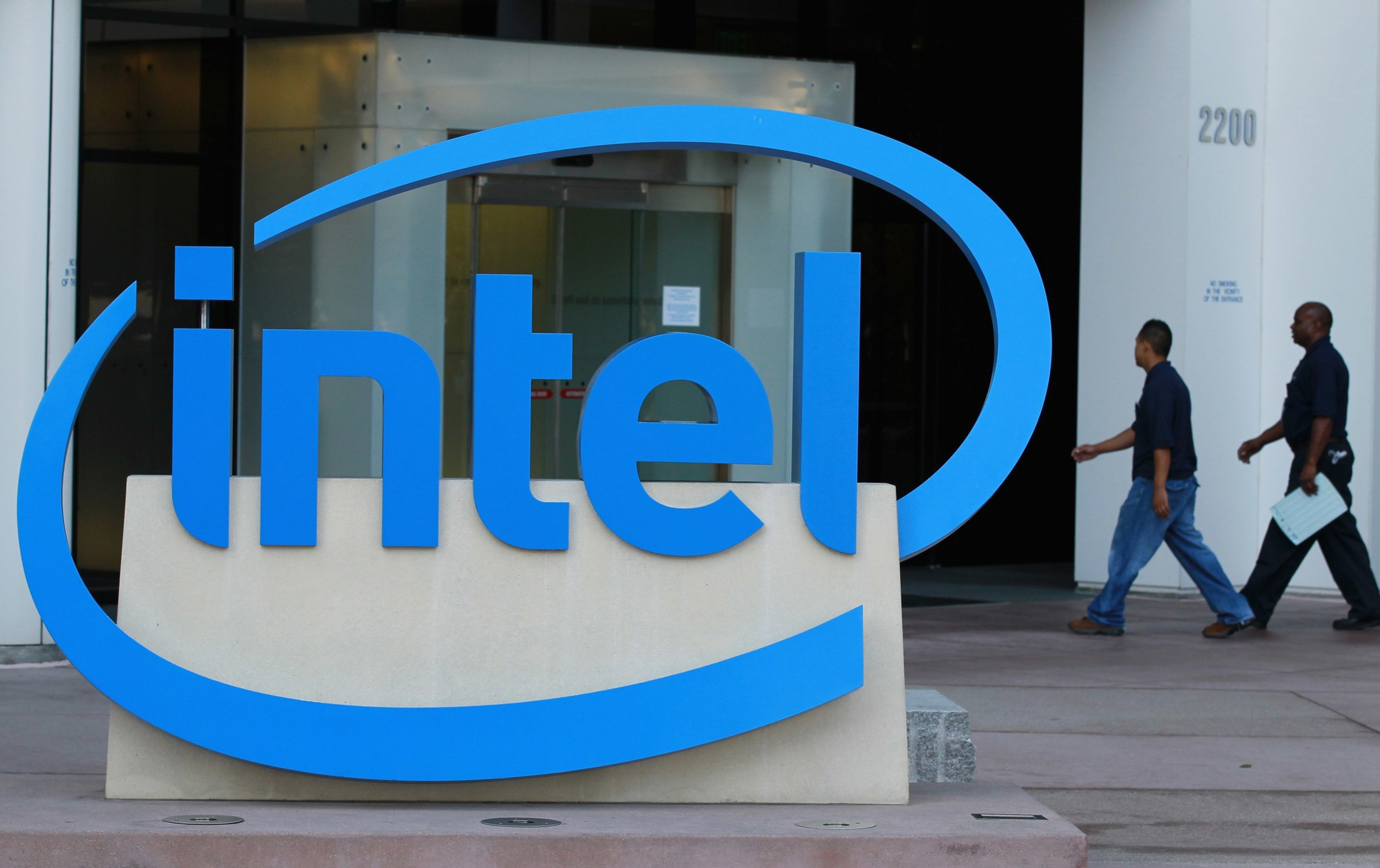 A large blue Intel logo is prominently displayed in front of a modern office building, with two people walking past it.