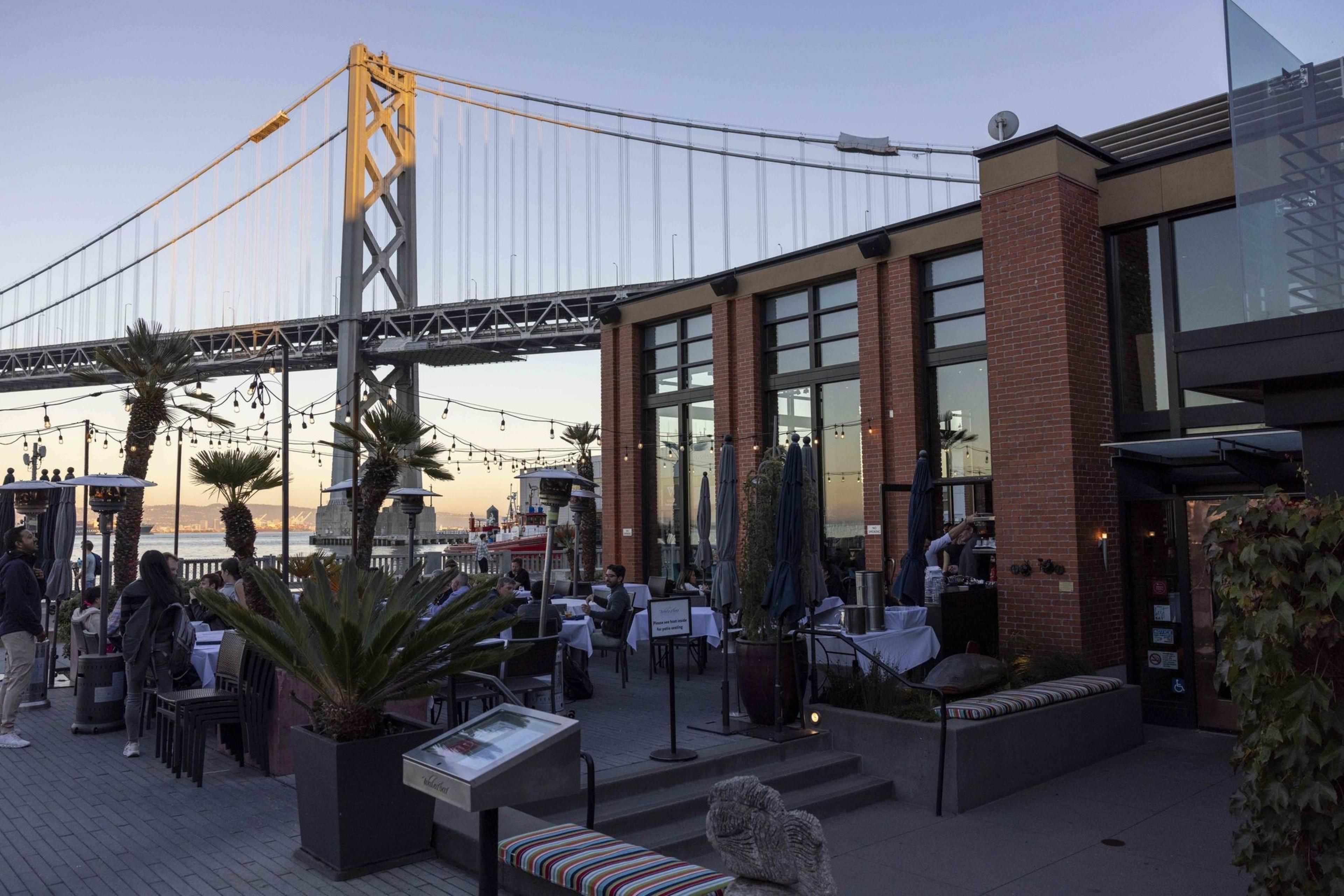 A vibrant outdoor restaurant by the waterfront features palm trees and string lights. A towering bridge spans the background. Diners sit at tables enjoying the view.