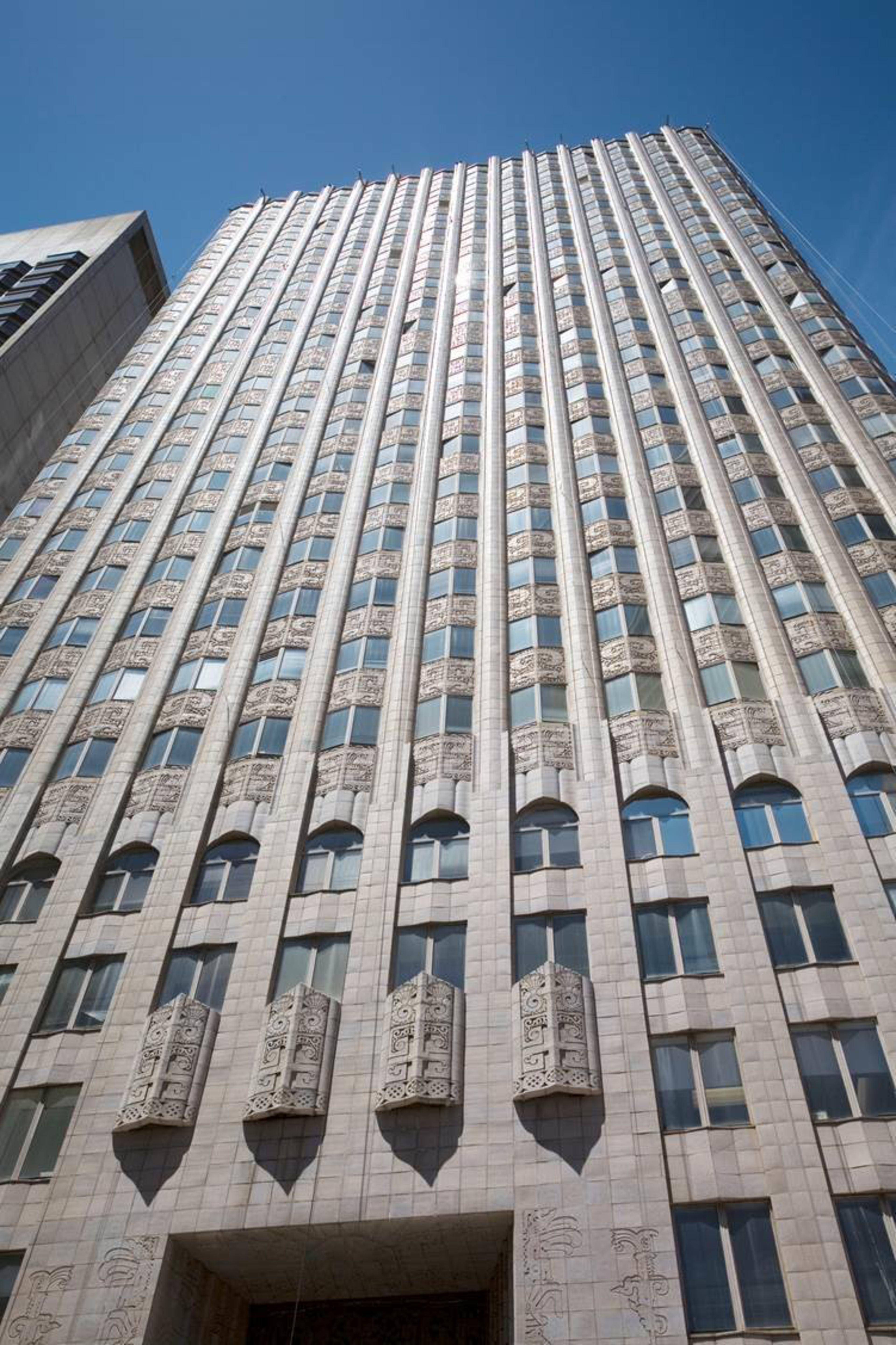 A tall building with vertical lines and ornate stone carvings rises against a clear blue sky, featuring numerous rectangular windows arranged uniformly.