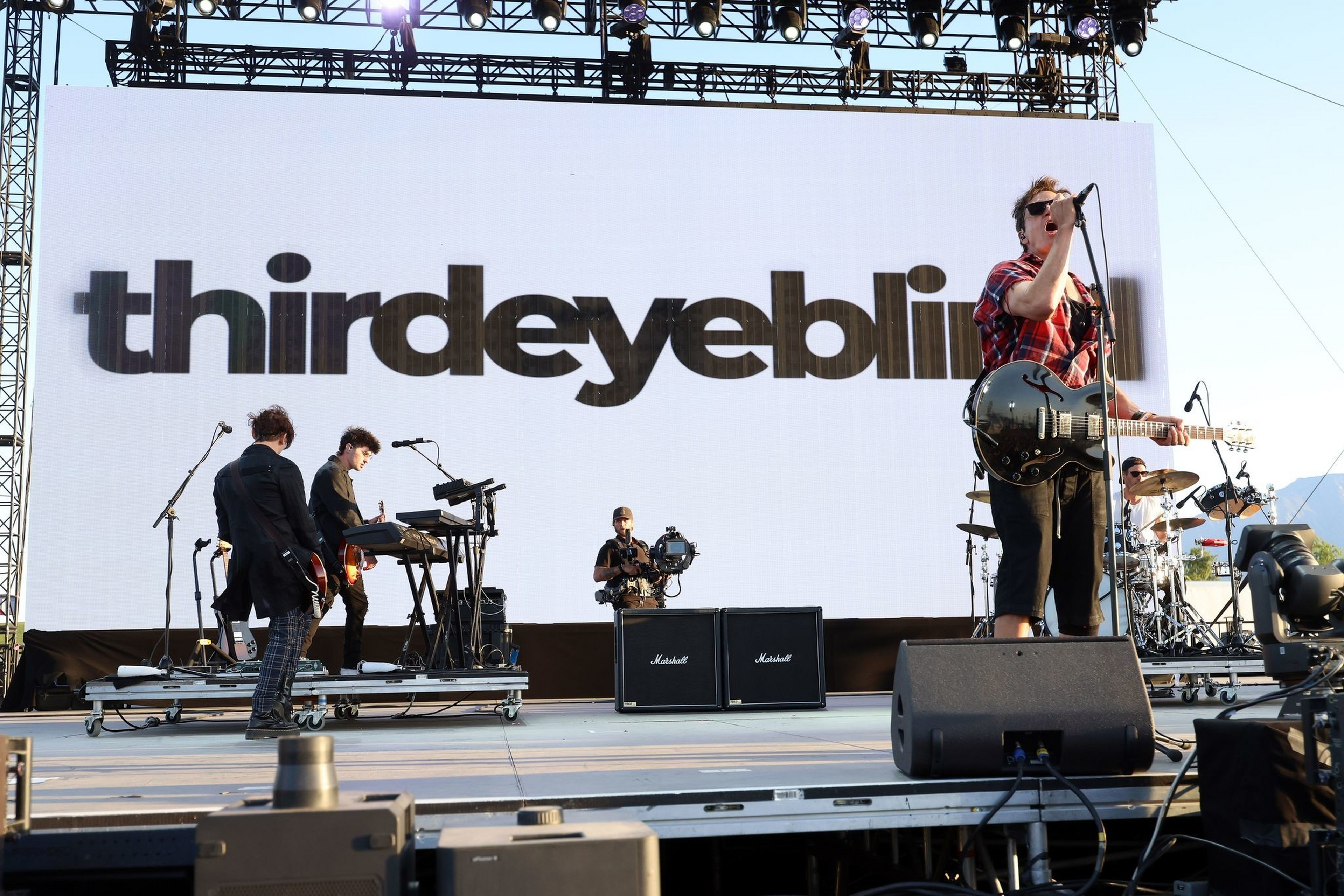 A band named “thirdeyeblin” performs on stage with members playing guitar, keyboard, and drums, backed by large speakers and a camera operator.