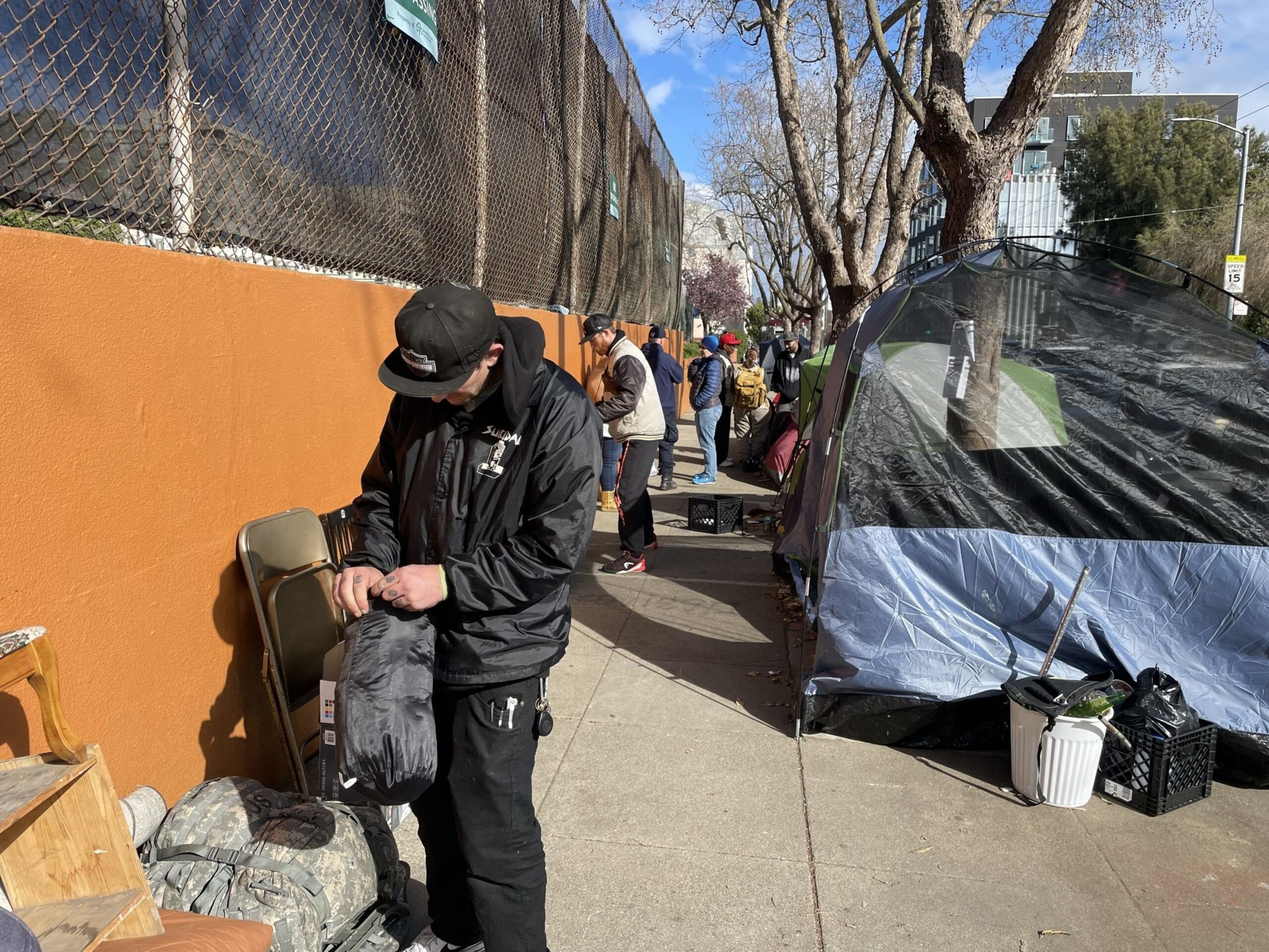 A man in black adjusts a black bag next to chairs and backpacks on a sidewalk, with tents, other people, and bare trees lining the street.