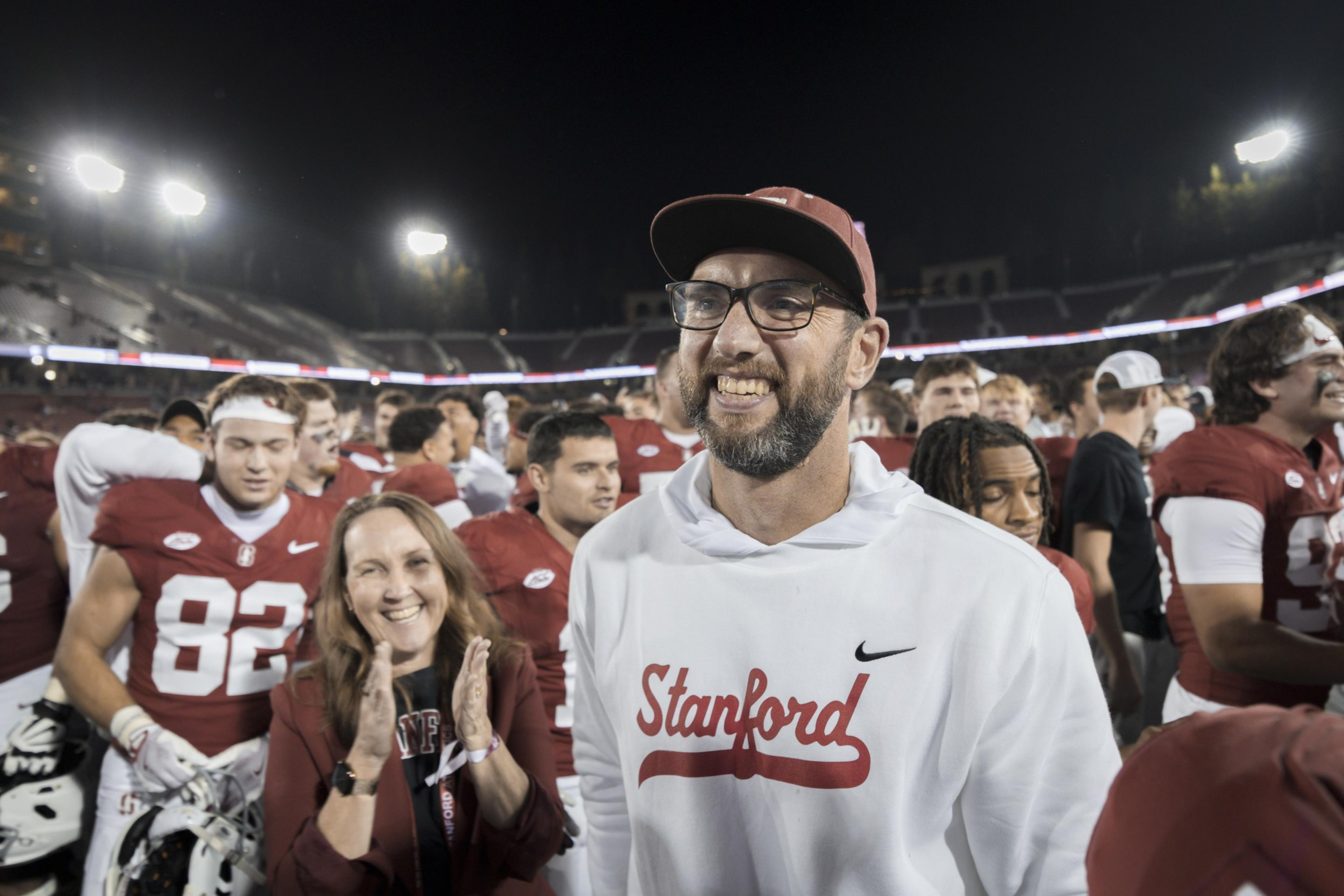A smiling man in a white Stanford hoodie and cap stands among cheering football players and a clapping woman on a lit stadium field at night.